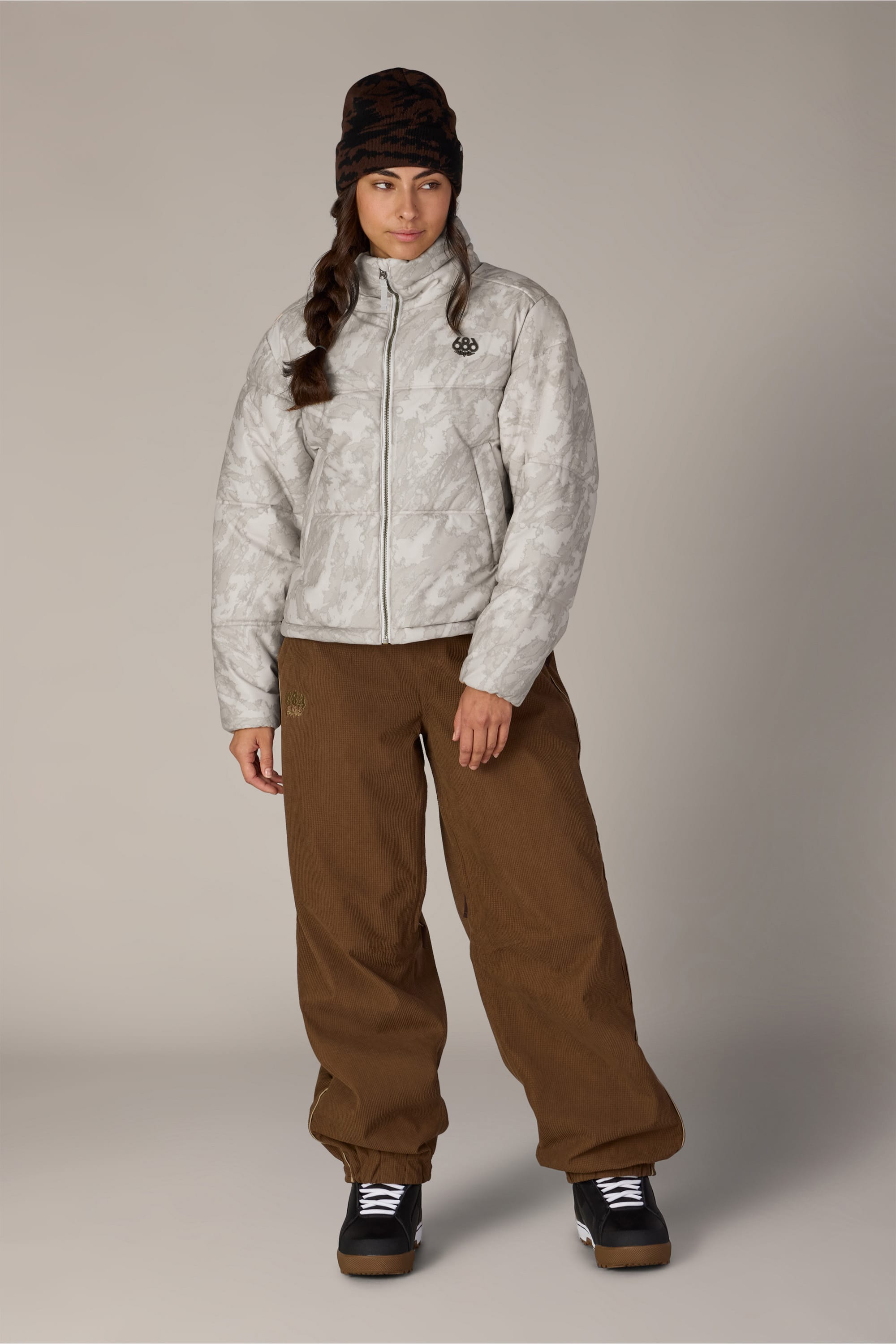 A woman wearing the 686 Women’s Gracious Puffer Jacket by 686, styled in a light gray pattern, pairs it with brown pants, black snow boots, and a black beanie as she stands against a plain light background, looking slightly to her left.