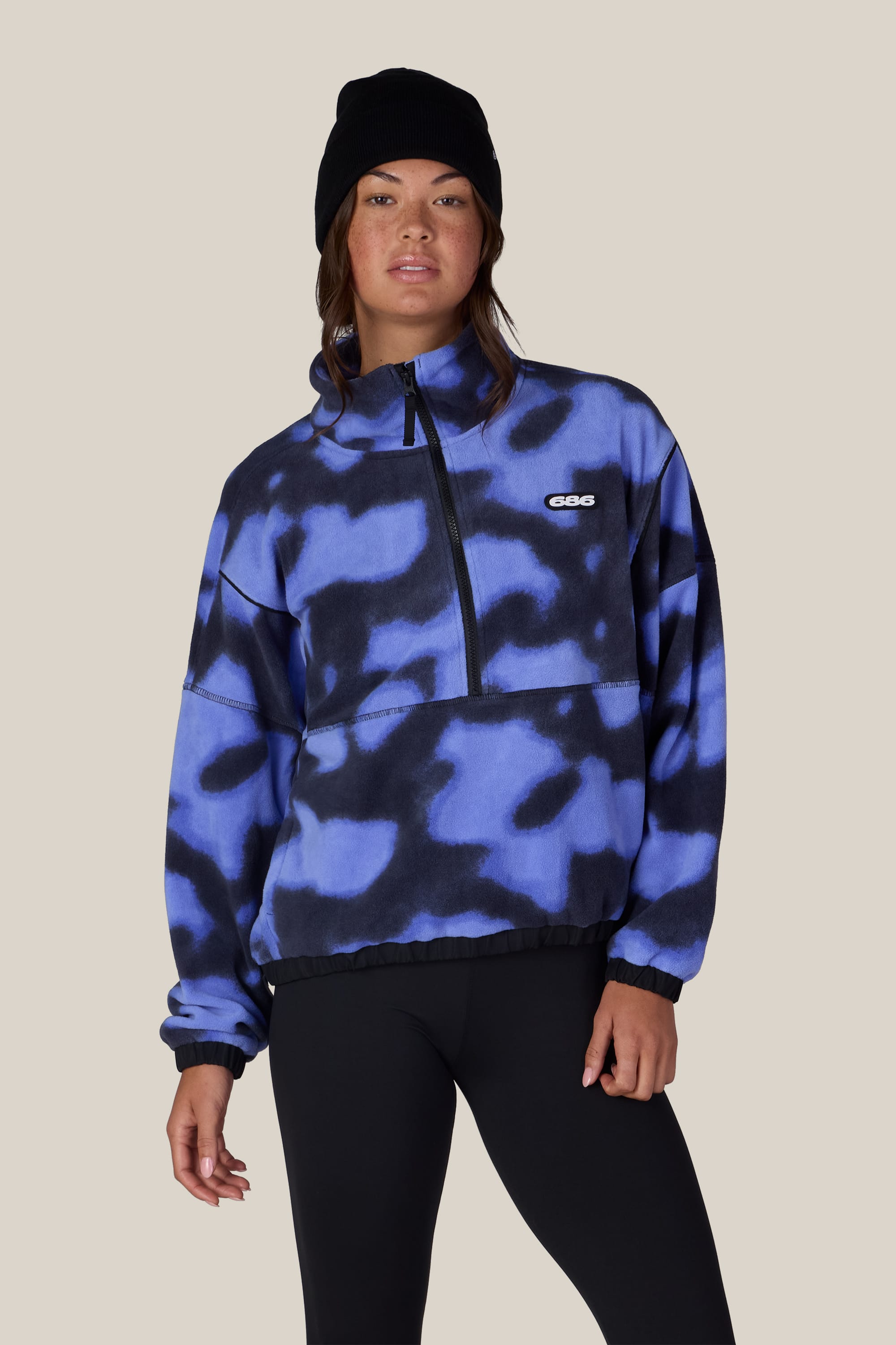 A person wearing a black beanie, black leggings, and the 686 Women’s Chimay Fleece Anorak—a blue and black patterned zip-up fleece pullover with a small logo—stands against a plain beige background, effortlessly showcasing the cozy versatility of this 686 jacket.
