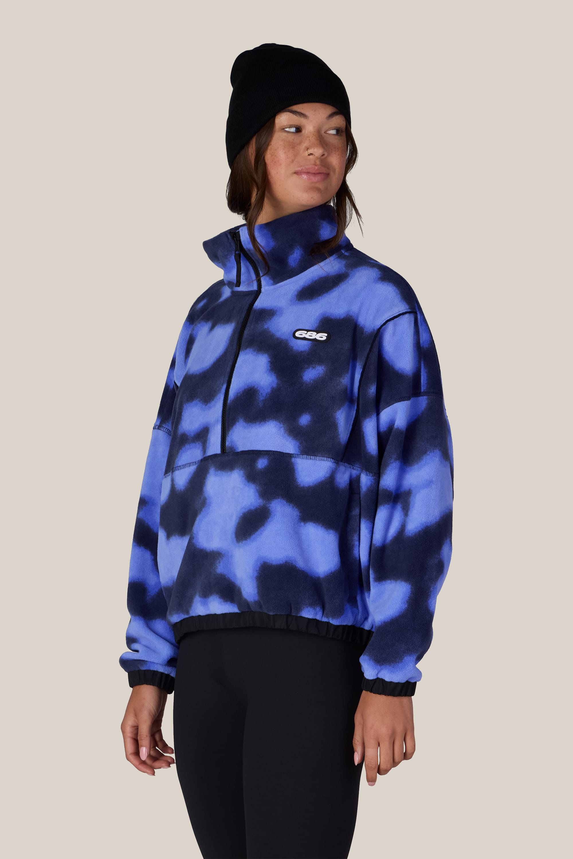 A person wearing a black beanie and the 686 Women’s Chimay Fleece Anorak—a blue and black patterned, zip-collar pullover from 686—stands against a plain beige background, showcasing the casual versatility of this 100% polyester jacket.
