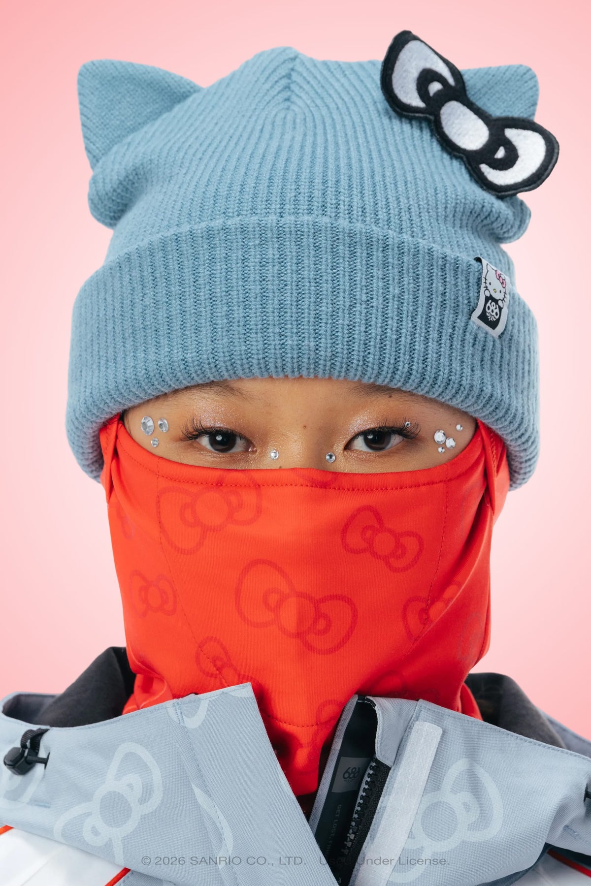 A person wearing the 686 Hurra Hinged Balaclava by 686, styled in blue with cat ears and a bow, paired with a gray jacket and rhinestone face decorations, stands against a pink background.