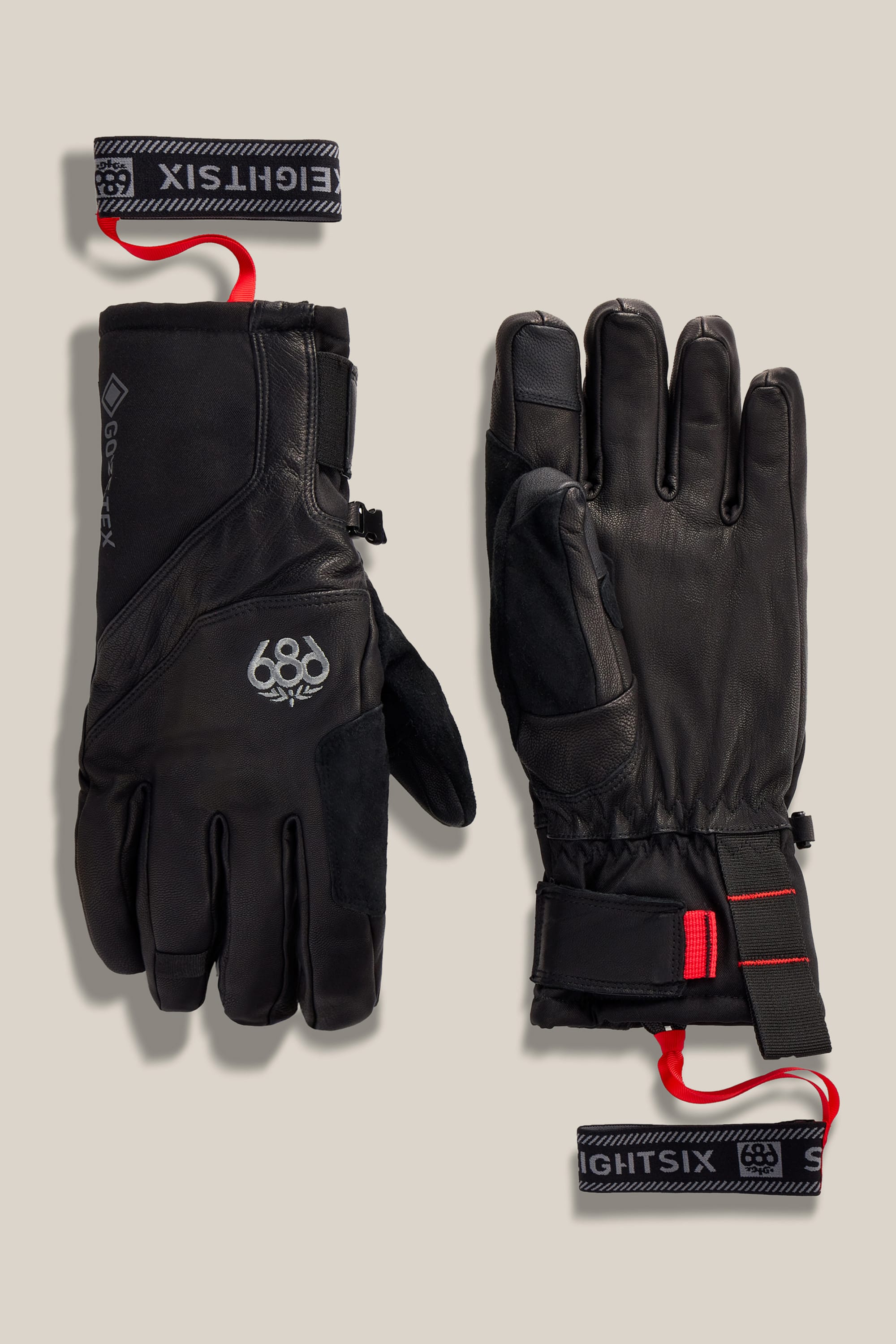 A pair of 686 Men’s GORE-TEX® Stronghold Leather Gloves, featuring black woven construction with red wrist straps and prominent 686 branding, displayed palm-up and palm-down on a beige background. Made from durable polyester spandex and finished with a supple leather palm for superior grip and comfort.