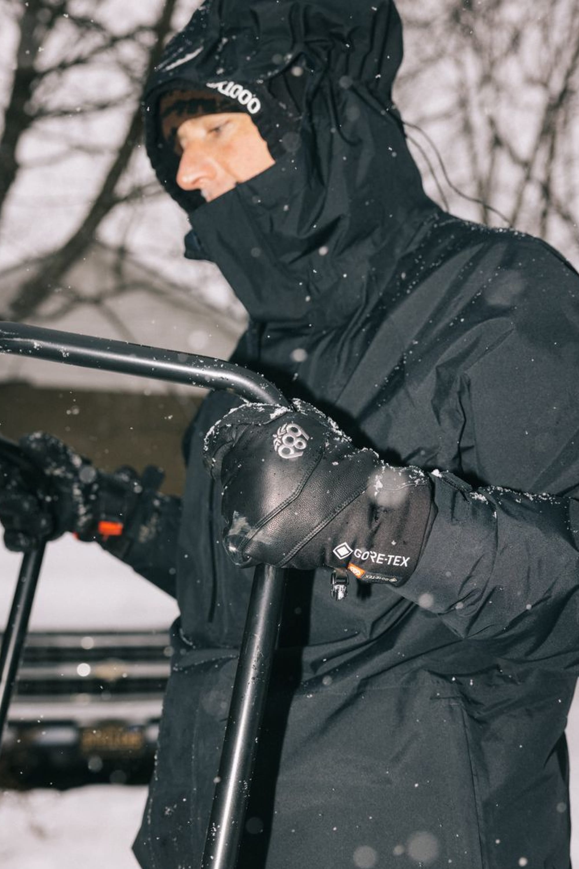 A person dressed in a black hooded winter jacket and wearing 686 Men’s GORE-TEX® Stronghold Leather Gloves by 686 holds metal bars outdoors in snowy weather, with blurred trees and a car in the background.