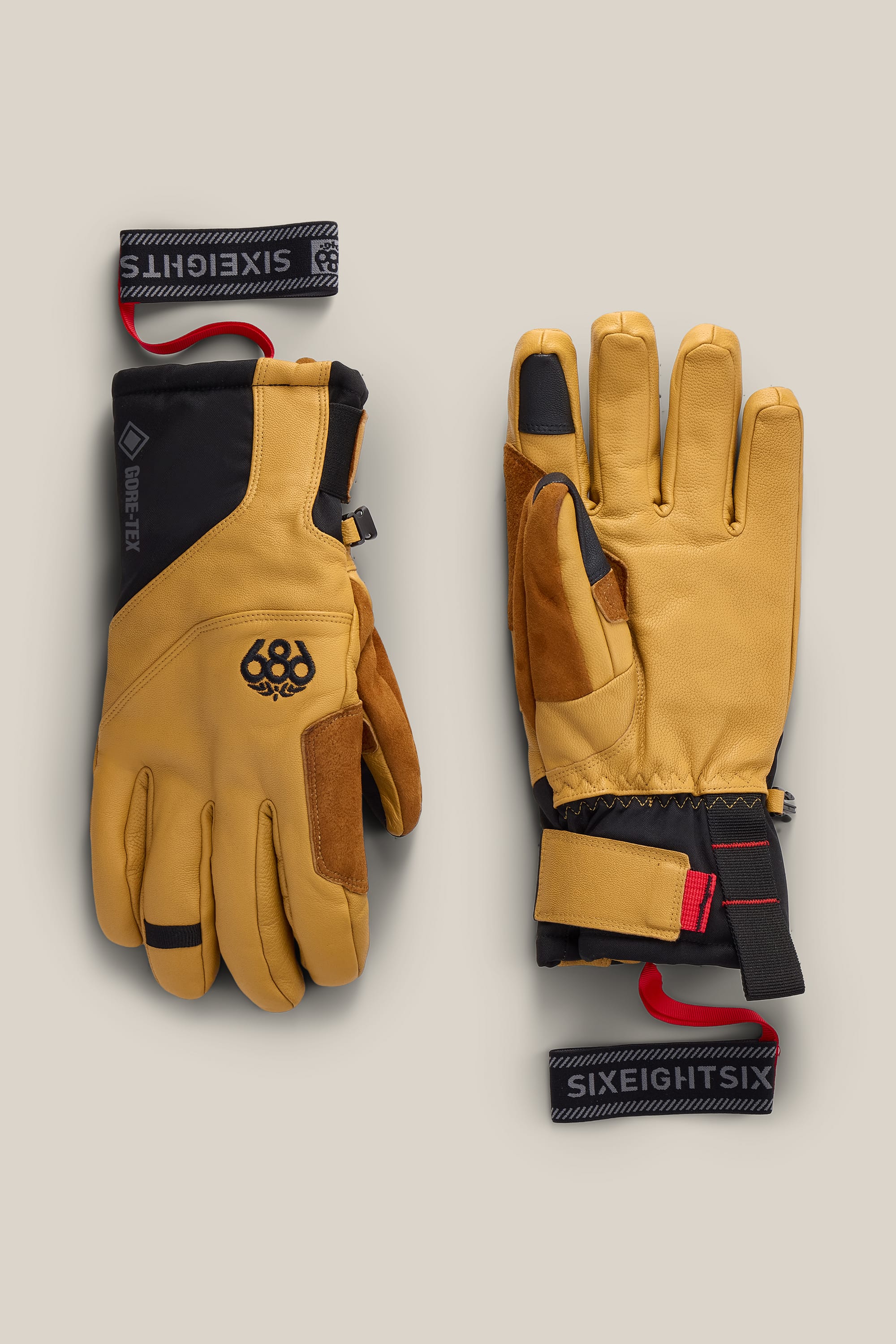 Two tan and black 686 Men’s GORE-TEX® Stronghold Leather Gloves with black wrist straps, each featuring reinforced fingertips, a durable leather palm for enhanced grip, red pull loops, and the 686 logo. One men’s woven glove is shown palm up and the other palm down.