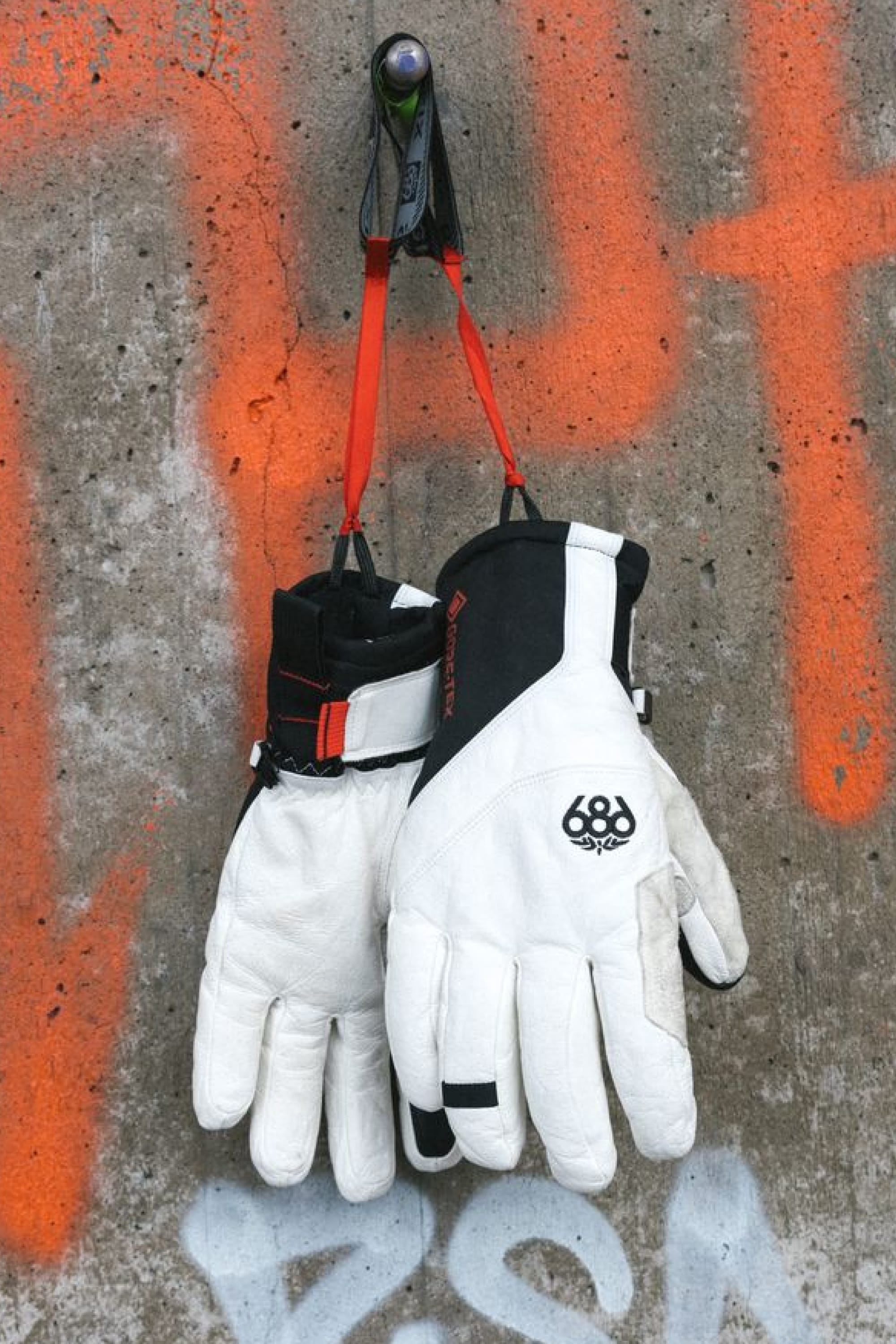 A pair of 686 Men’s GORE-TEX® Stronghold Leather Gloves with leather palms and black accents hangs by red straps from a hook on a concrete wall adorned with orange graffiti.