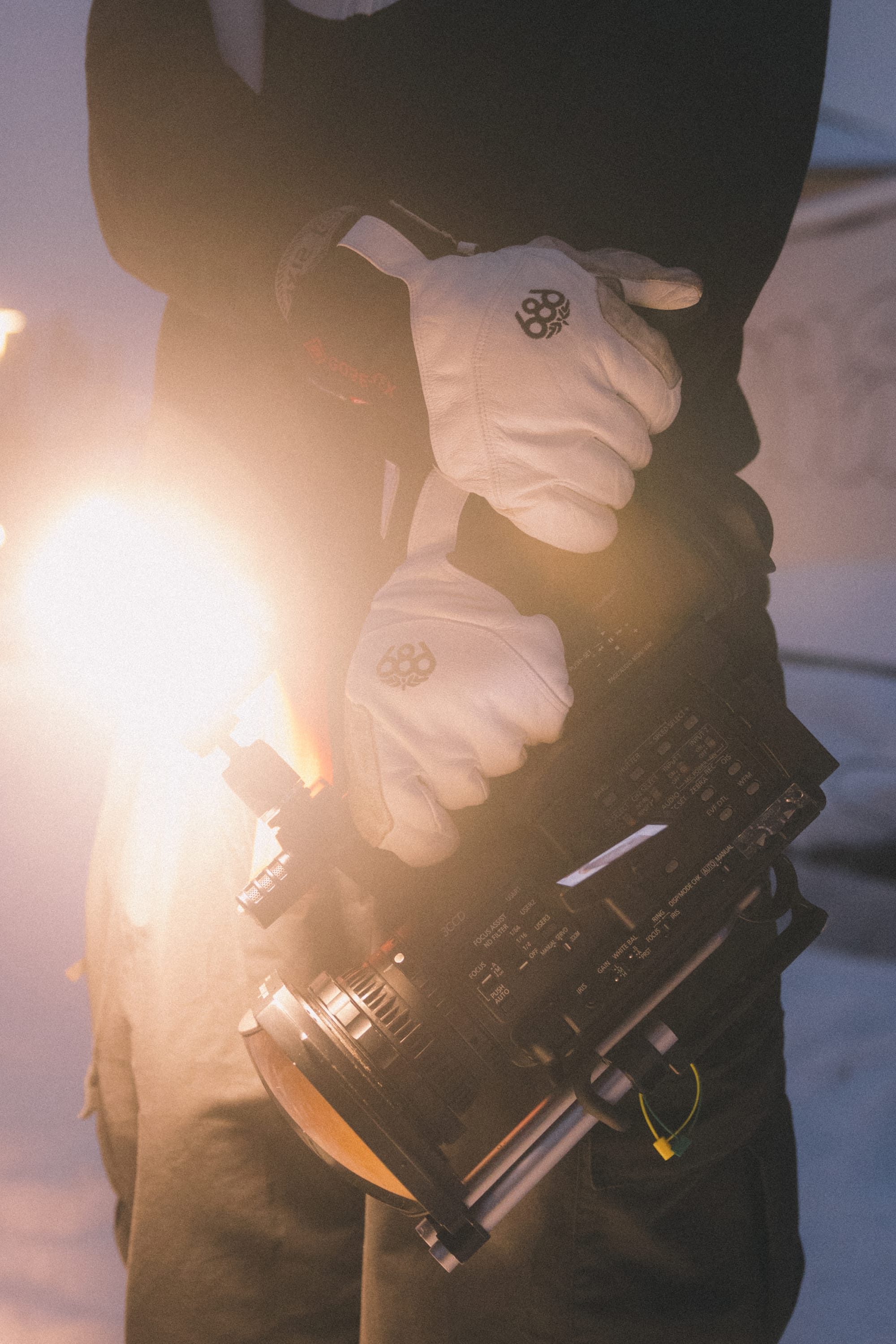 Wearing the 686 Men’s GORE-TEX® Stronghold Leather Glove by 686, a person stands outdoors in a muted, wintry setting holding a professional video camera with a bright light attached.