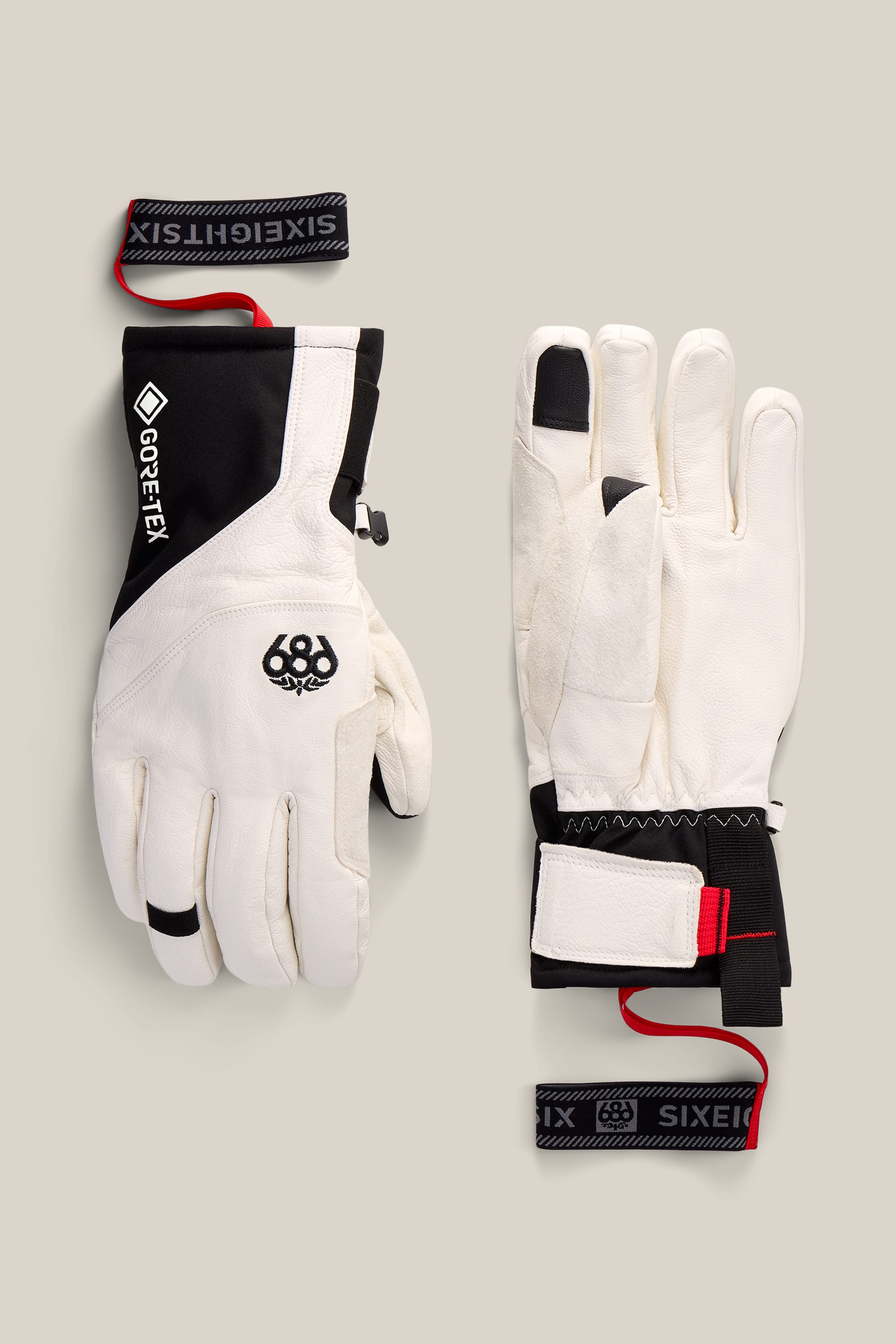 A pair of 686 Men’s GORE-TEX® Stronghold Leather Gloves in black and white, featuring insulated construction, durable leather palms, and red-and-black wrist straps from the brand 686, are displayed palm up and palm down on a light beige background.