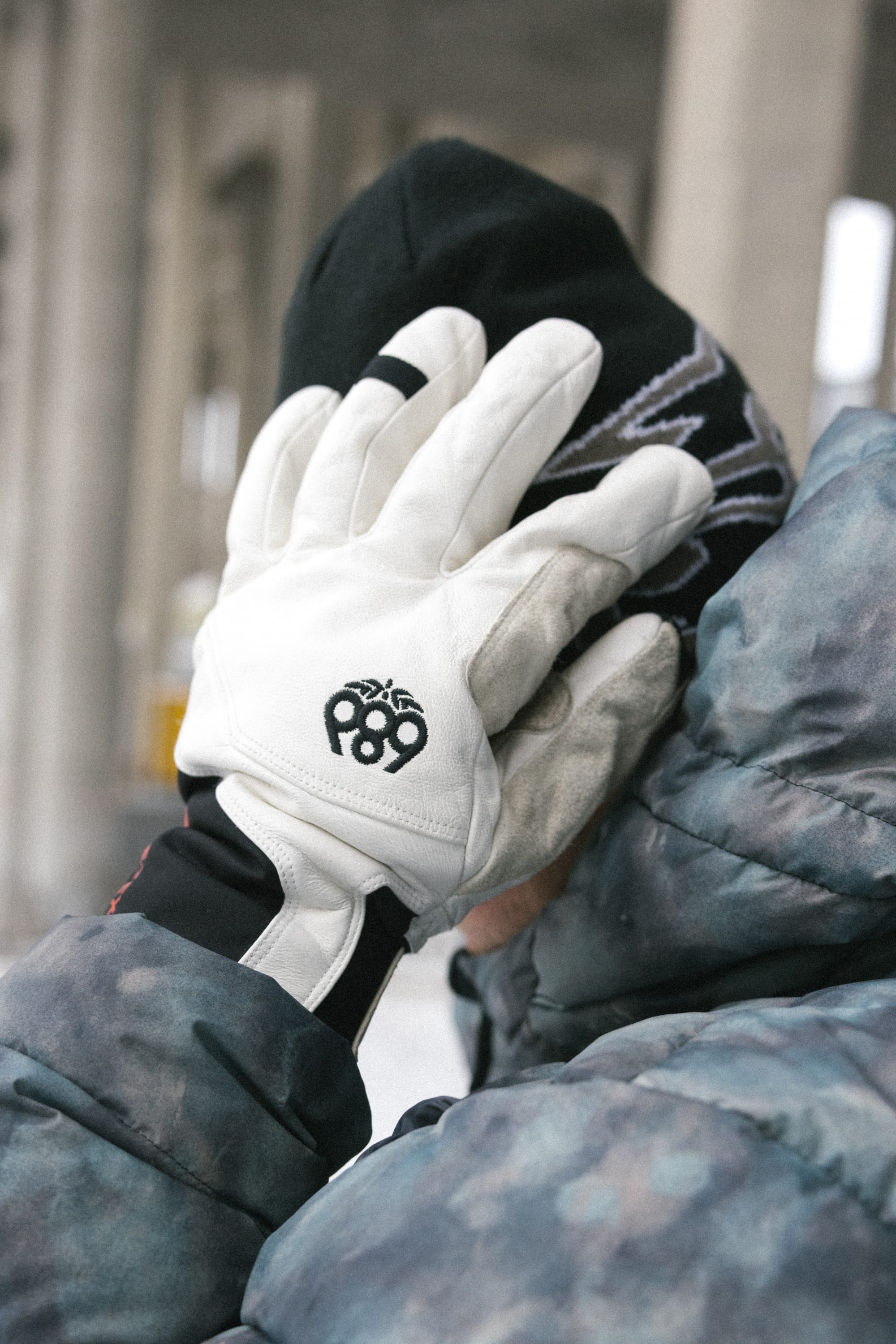 A person in a winter jacket and knit hat covers their face with a gloved hand. The white 686 Men’s GORE-TEX® Stronghold Leather Glove, featuring a black “89” logo, is crafted from polyester spandex with a durable leather palm. The background is blurred, suggesting an indoor or urban setting.