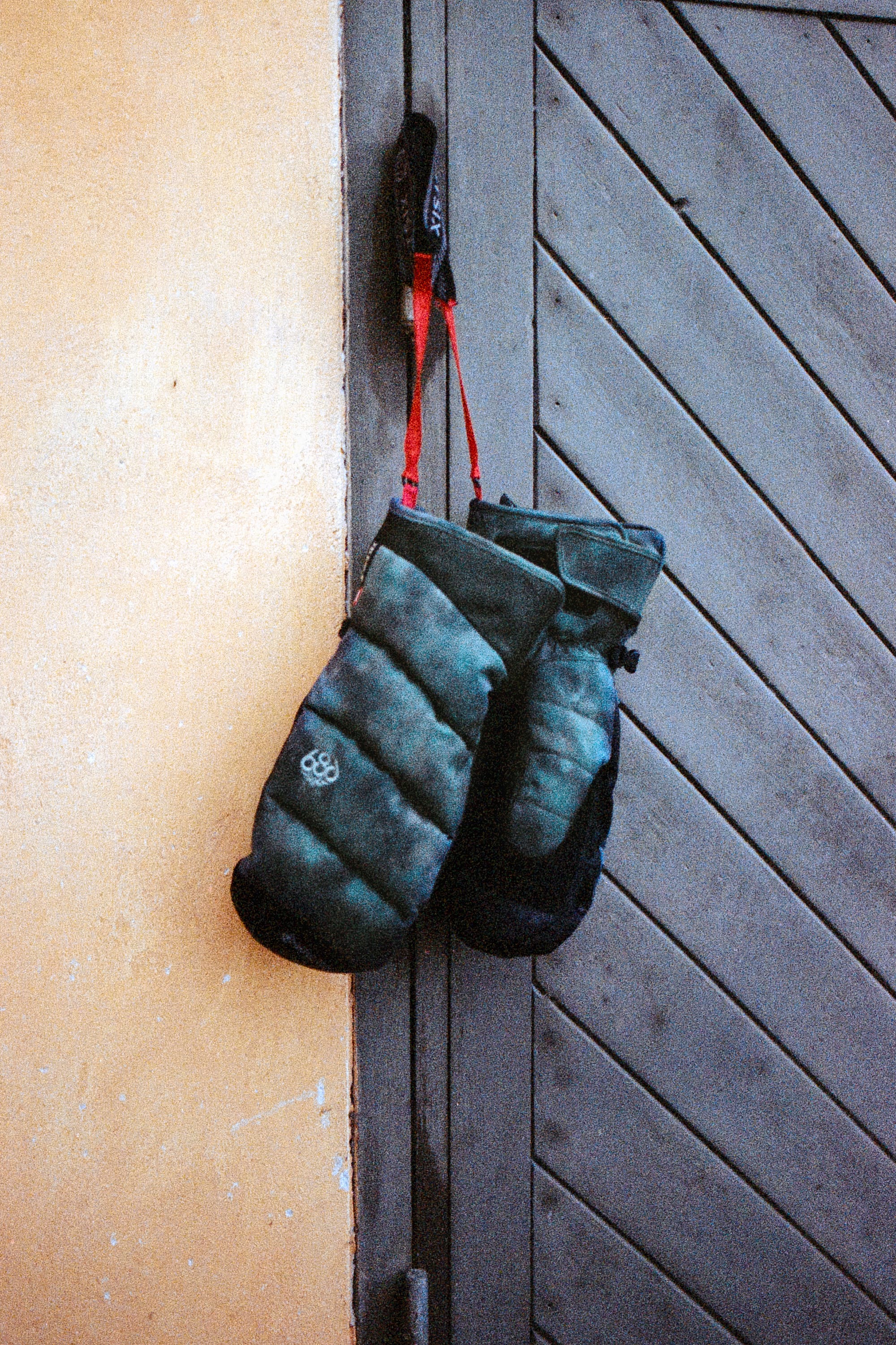 A pair of Men’s GORE-TEX® Buffer Insulated Mitts by 686 in dark green with red straps hang on a hook against a yellow wall and a gray wooden door with diagonal panels.