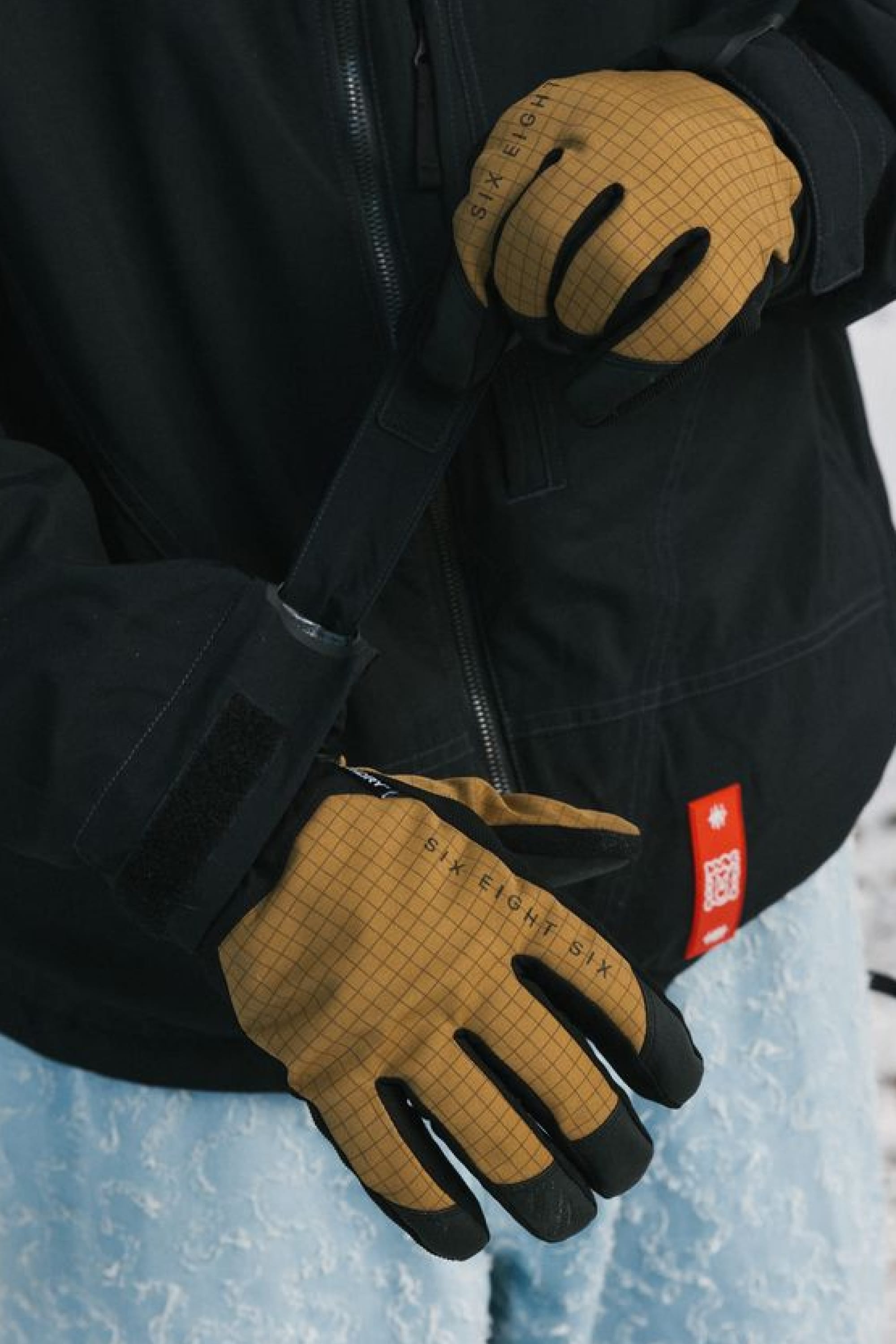 A person wearing 686 Men’s Crush Insulated Gloves in a light brown grid pattern and a black jacket adjusts a strap, with blue patterned pants partially visible.