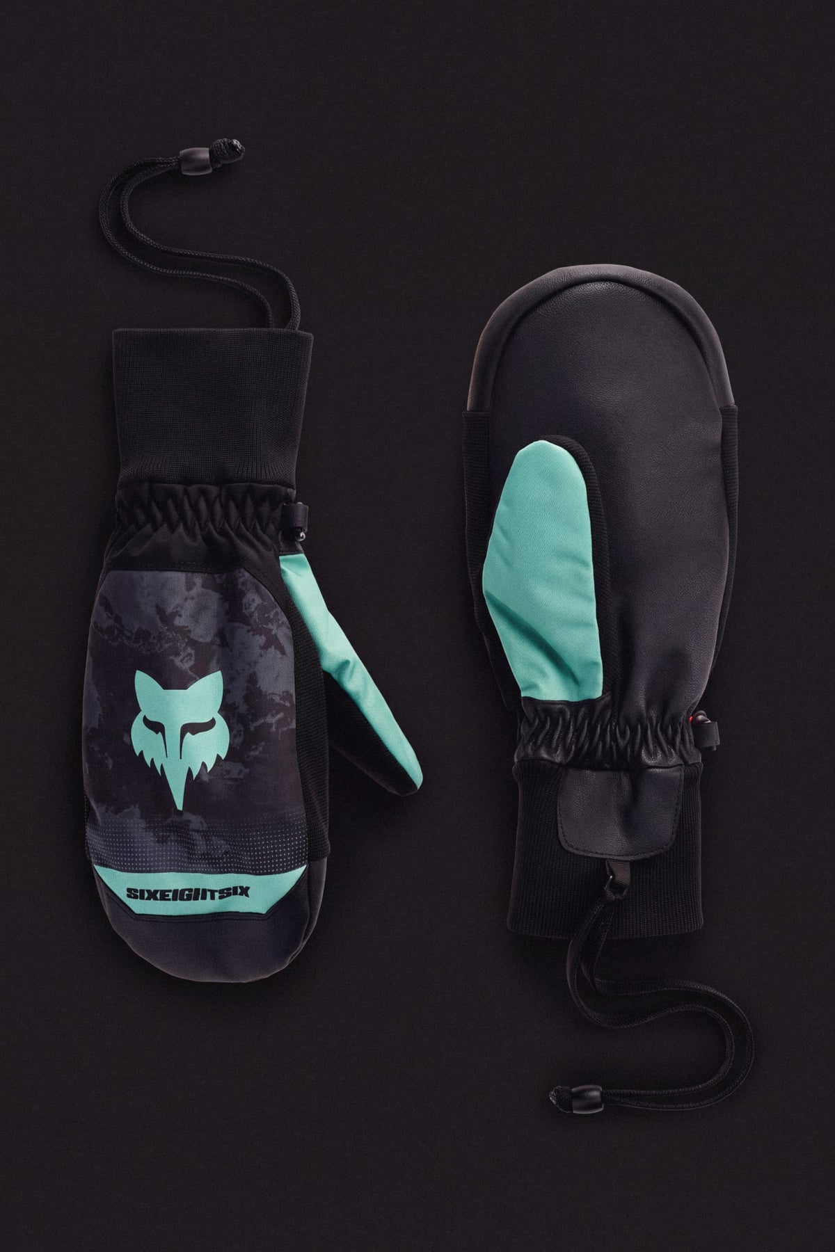 The 686 Men’s Crush Insulated Mitt by 686, shown in black with teal accents and a fox logo, features wrist straps and elastic wrists; one mitten displays the palm, the other the back on a black background.