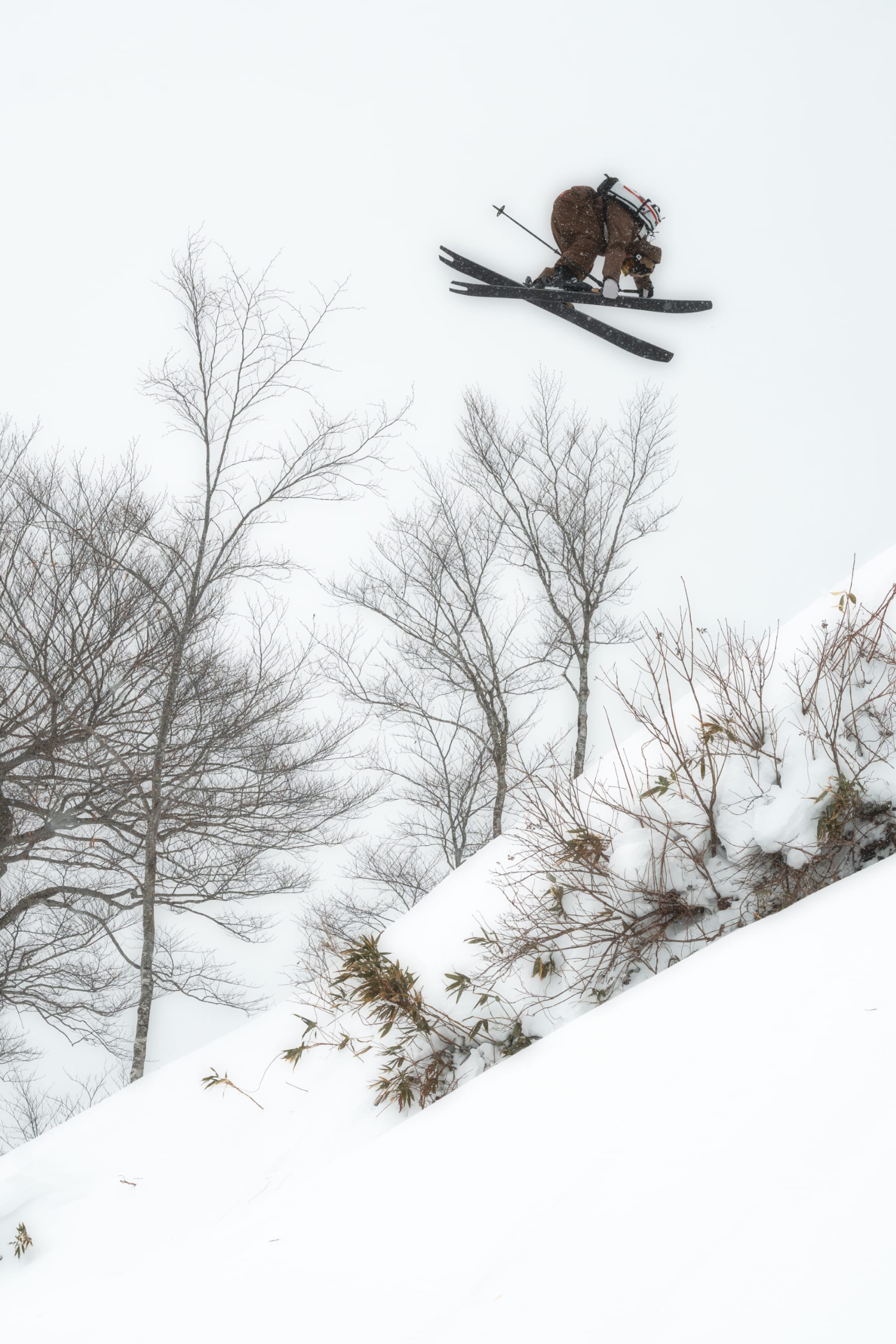 A skier in brown gear is captured mid-air above a snowy slope, wearing the 686 Men’s Crush Insulated Mitts as he soars past leafless trees and over the snow-covered ground.
