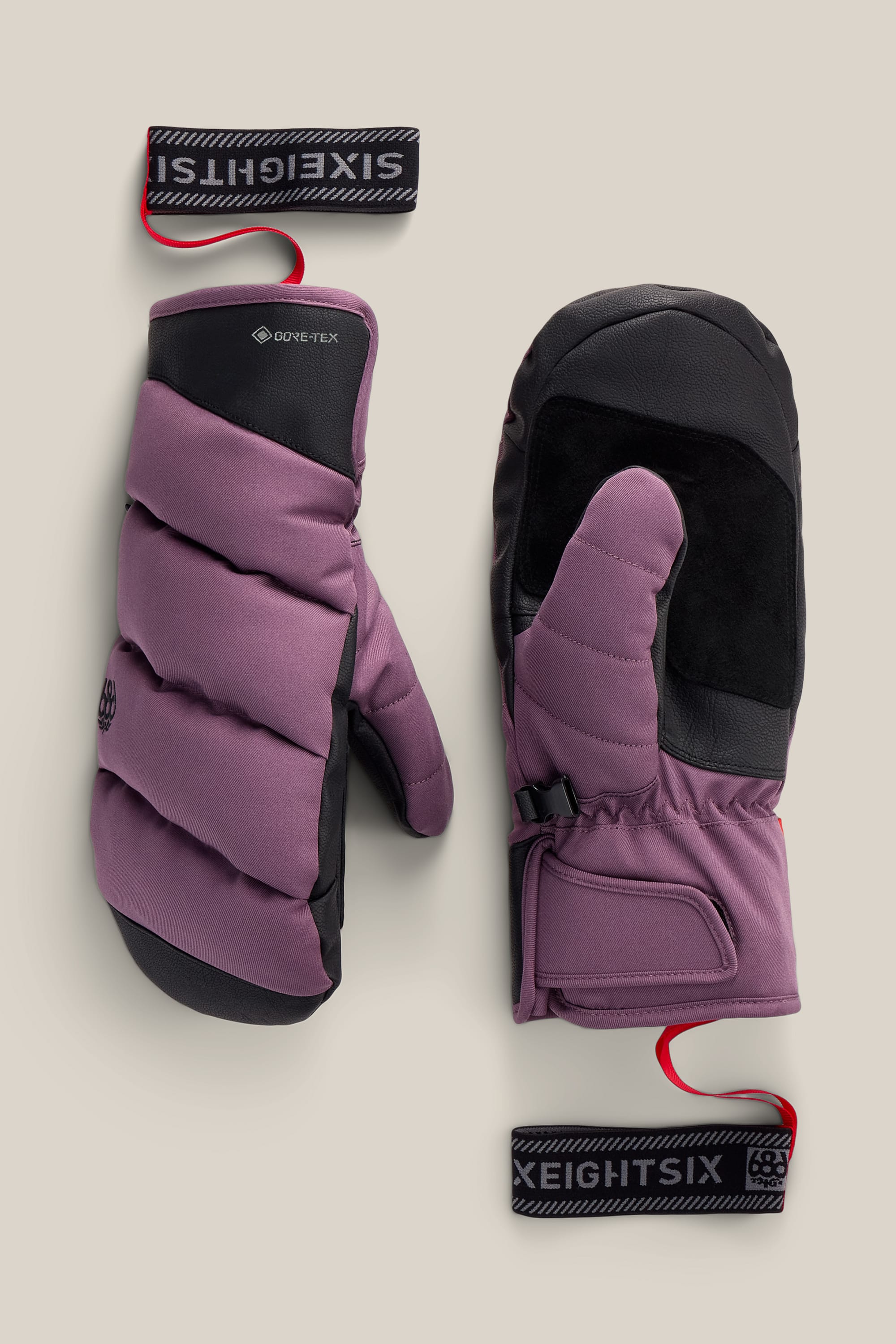 A pair of 686 Women’s GORE-TEX® Buffer Insulated Mitts in purple and black, featuring black palm patches, red wrist straps, and detachable logo tags reading SIXEIGHTSIX. The mittens are displayed flat on a beige background.