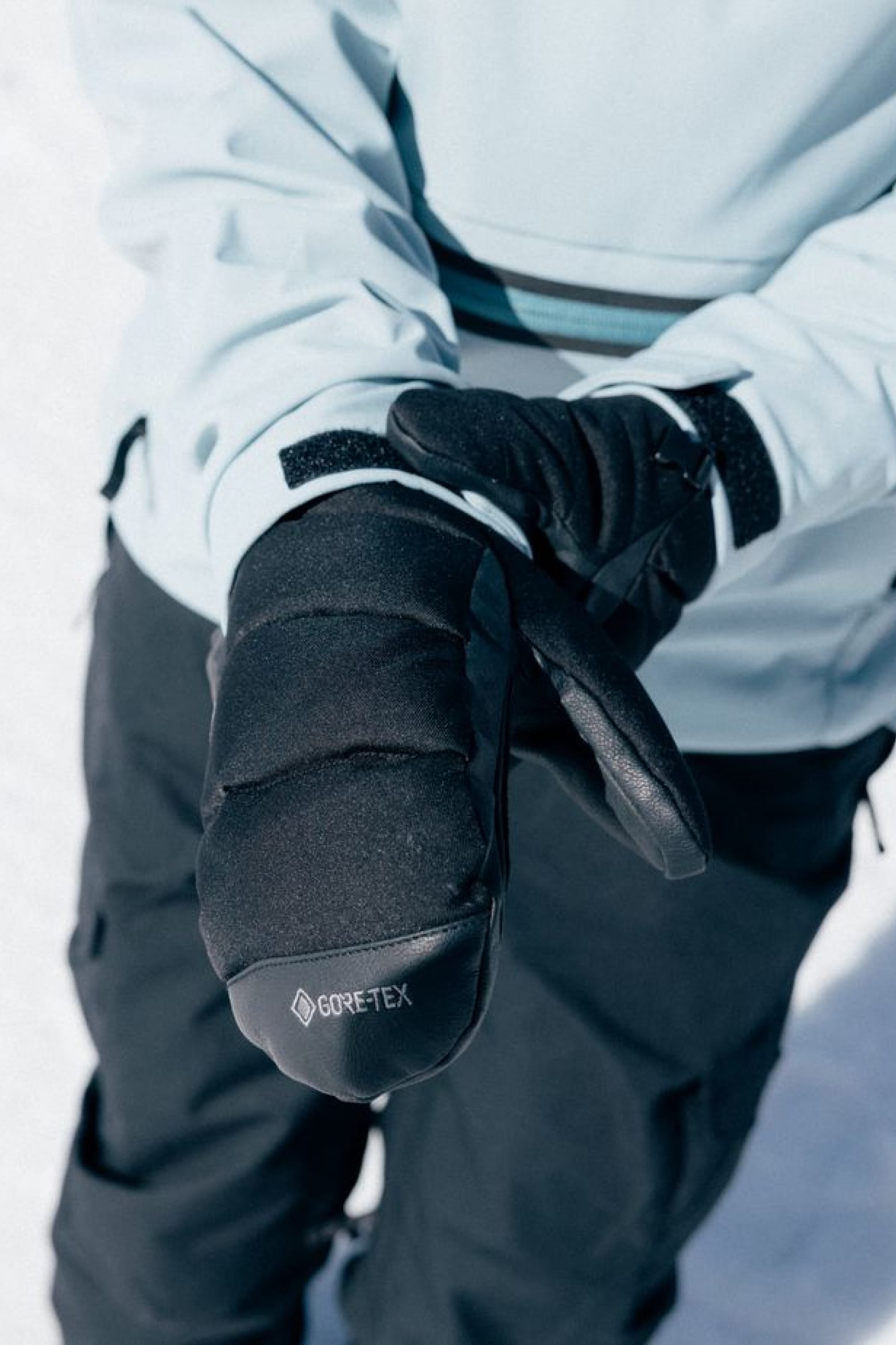 A person wearing black 686 Women’s GORE-TEX® Buffer Insulated Mitts and a light blue jacket stands on snow, adjusting one mitt with the other hand.