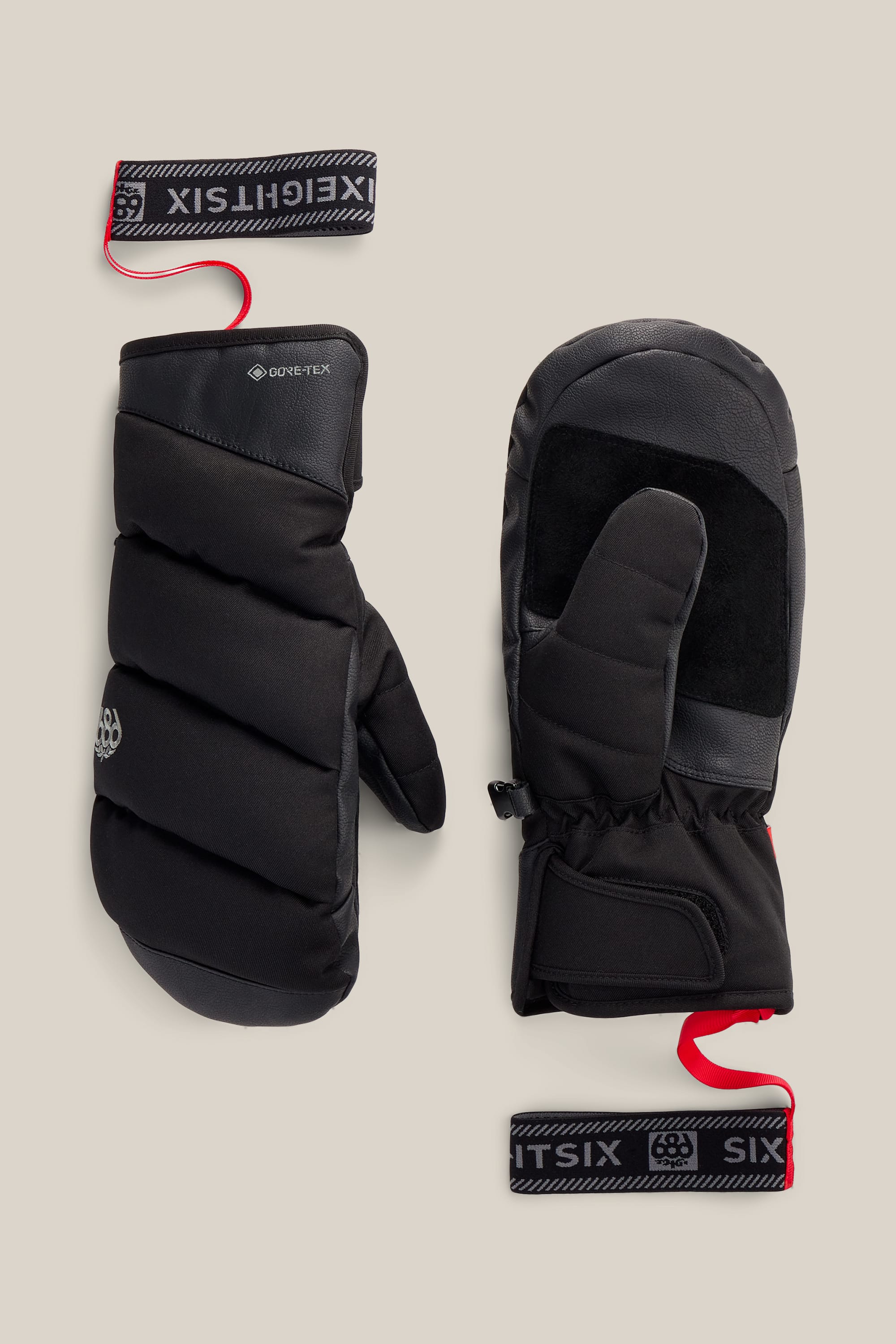 A pair of black Women’s GORE-TEX® Buffer Insulated Mitts by 686, featuring leather palms and thumb patches, is shown front and back. Each mitt displays a red EIGHT SIX wrist strap, all set against a beige background.