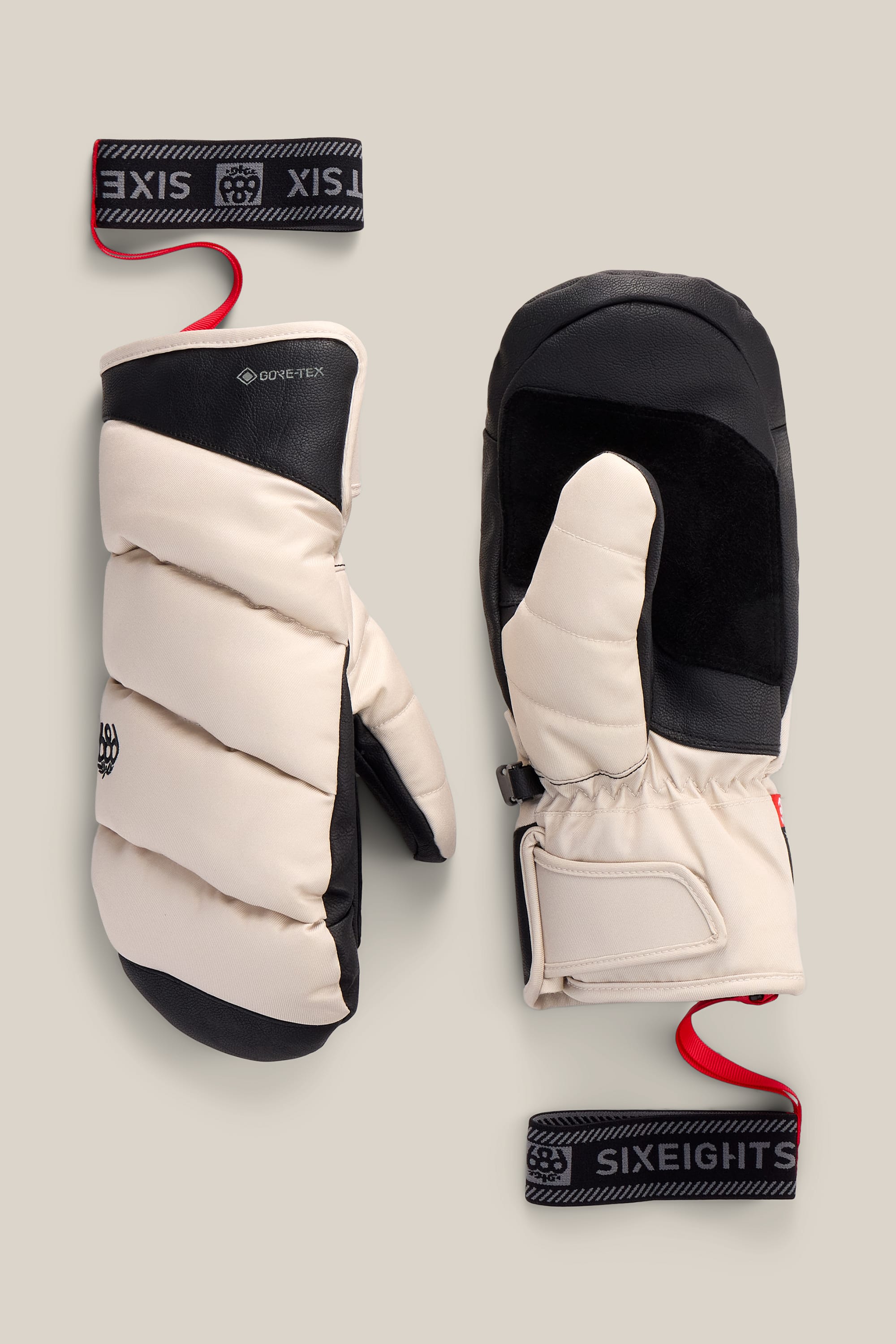 A pair of Women’s GORE-TEX® Buffer Insulated Mitts by 686 in beige and black, featuring padded construction, Velcro straps with red pull tabs, and the 686 logo printed on the black and red wrist straps. Displayed on a neutral background, these stylish poly mitts offer both comfort and distinctive design.