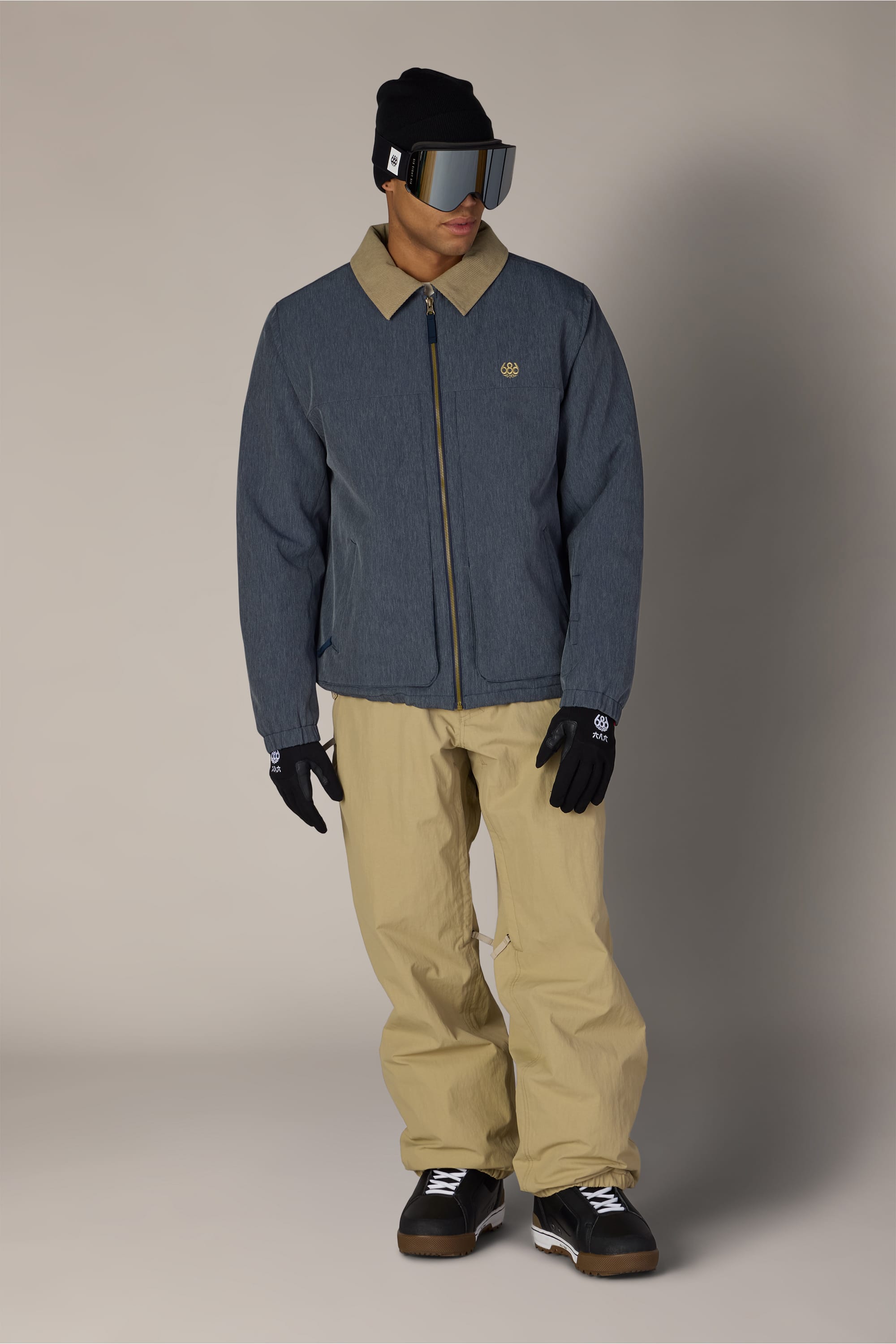 A person wearing the 686 Men’s 686 Camber Insulated Jacket by 686 in blue with a tan collar, tan pants, black gloves, black winter boots, a black beanie, and reflective ski goggles stands against a plain background.
