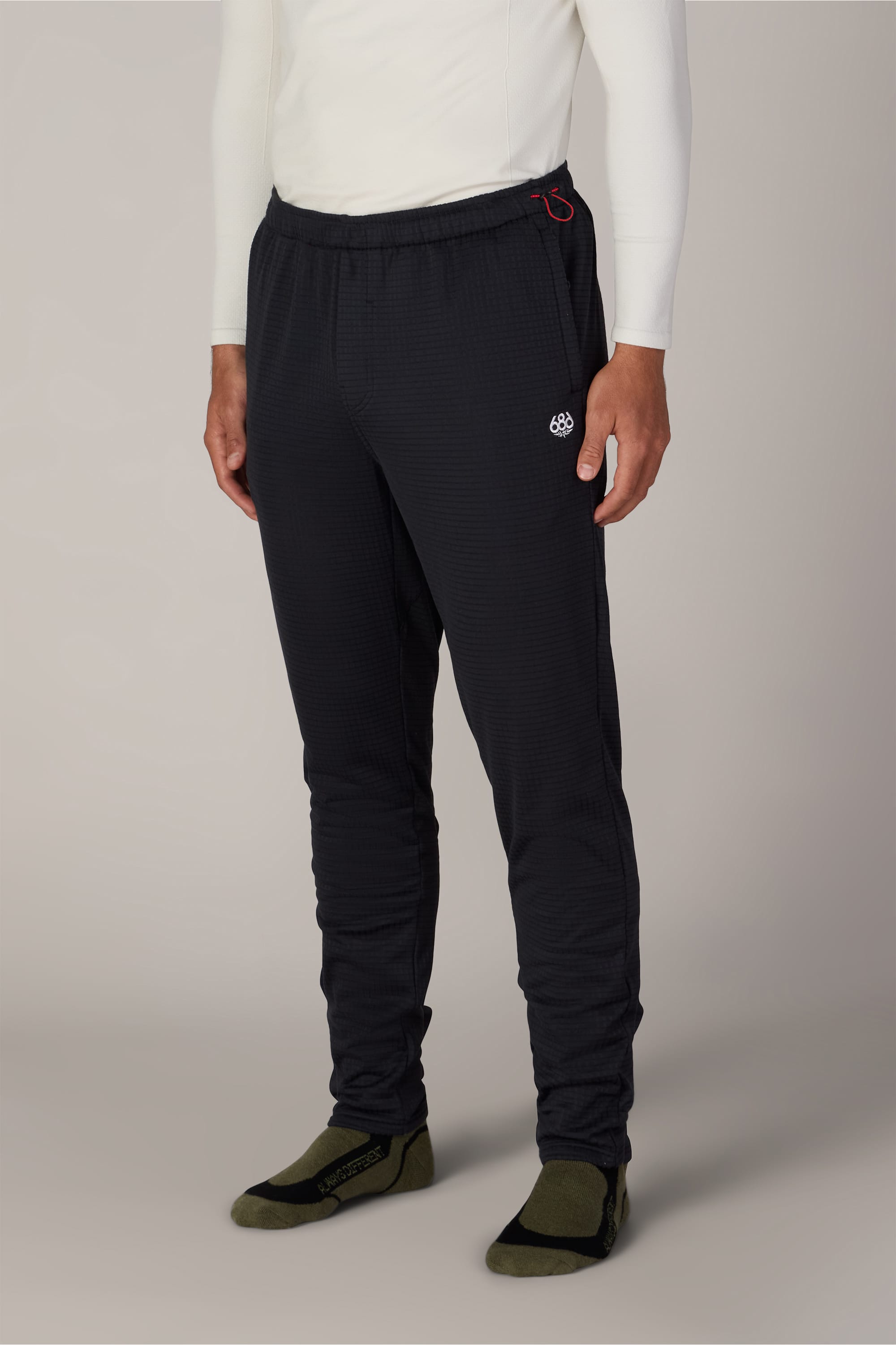 A person wearing a white long-sleeve shirt, 686 Men’s Mission Grid™ Pant by 686 in a dark knit with a small logo, and olive green slip-on shoes, standing against a neutral background.