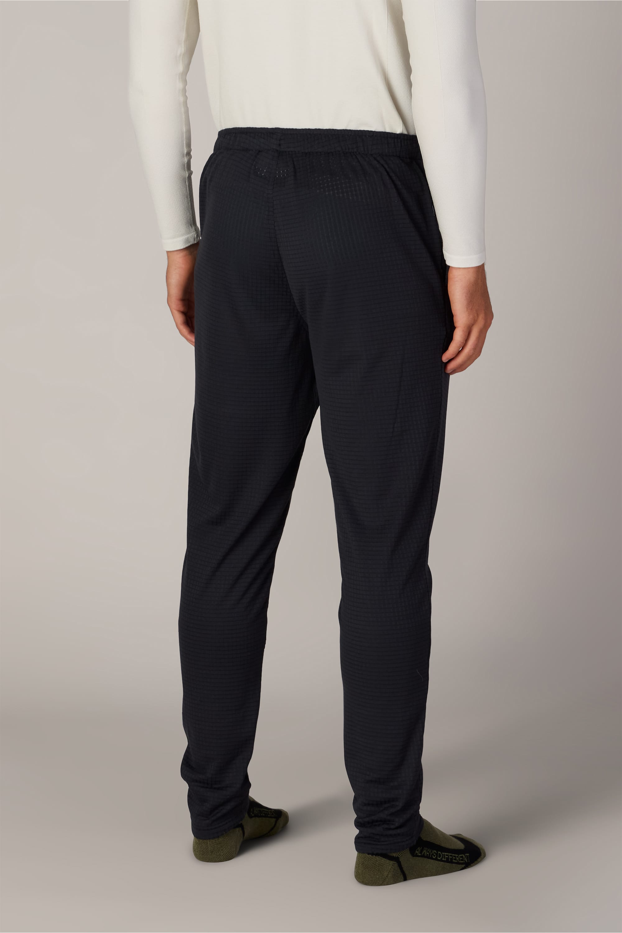 A person stands facing away, wearing a long-sleeve white shirt, dark socks, and the 686 Men’s Mission Grid™ Pant by 686—a pair of textured, dark men’s knit pants. The background is plain and neutral.