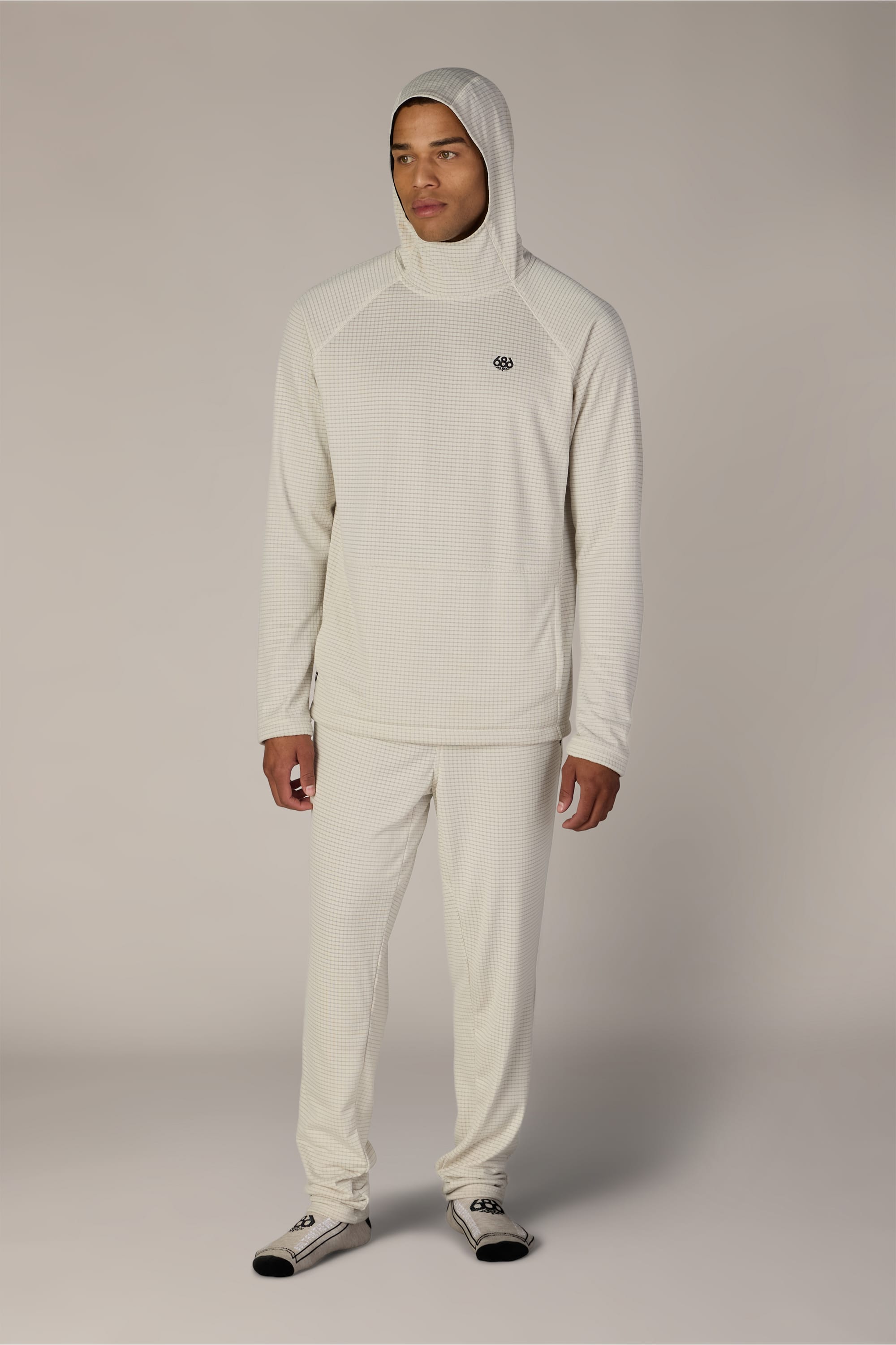 A person stands facing forward, wearing a light beige hooded sweatshirt and the 686 Men’s Mission Grid™ Pant by 686, which features a textured knit pattern. They also have on gray socks, with a plain, neutral background.