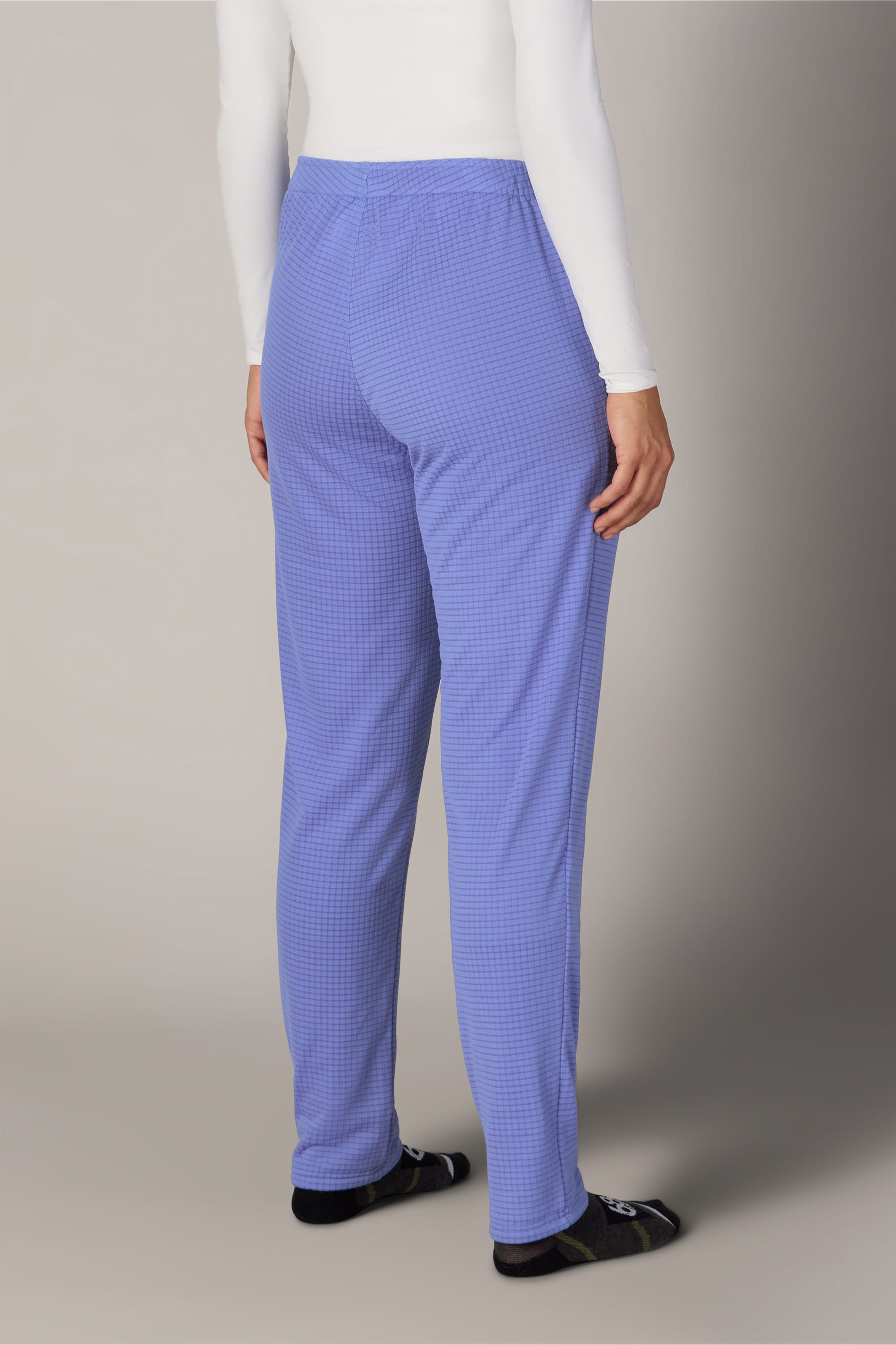 A person stands facing away, wearing a white long-sleeve shirt, dark socks, and light blue checkered 686 Women’s Mission Grid™ Pant by 686. The background is plain and neutral.