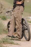 A person in 686 Mens Platform Bike Pant - Relaxed Fit stands by a mountain bike on a dirt path, one hand on their hip. The scene includes greenery and blurred hills in the background.