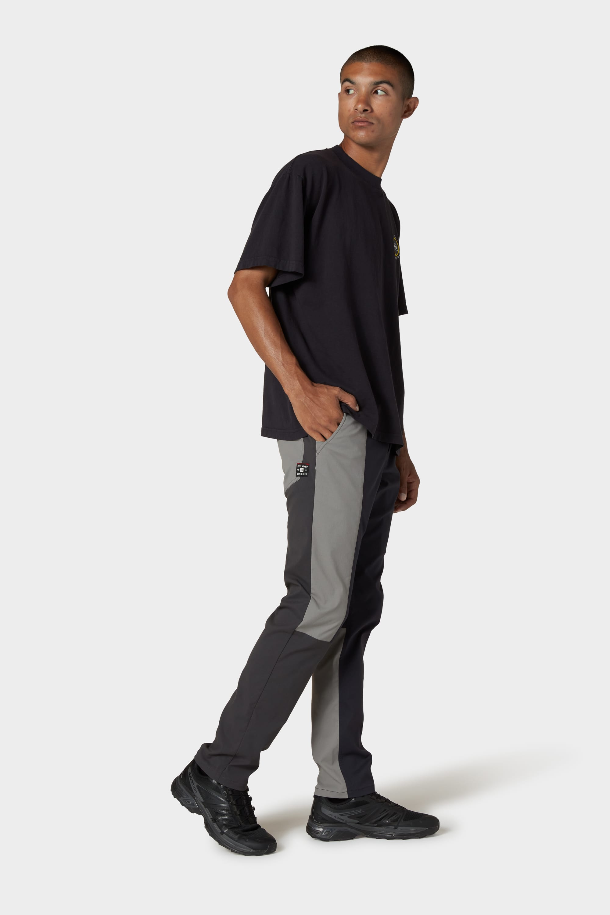 A person stands against a plain background, wearing a black t-shirt, black sneakers, and the 686 Men's Everywhere® Reclaimed Pant - Slim Fit by 686, made from deadstock fabrics, with one hand in their pocket and looking to the side, reflecting sustainable fashion.