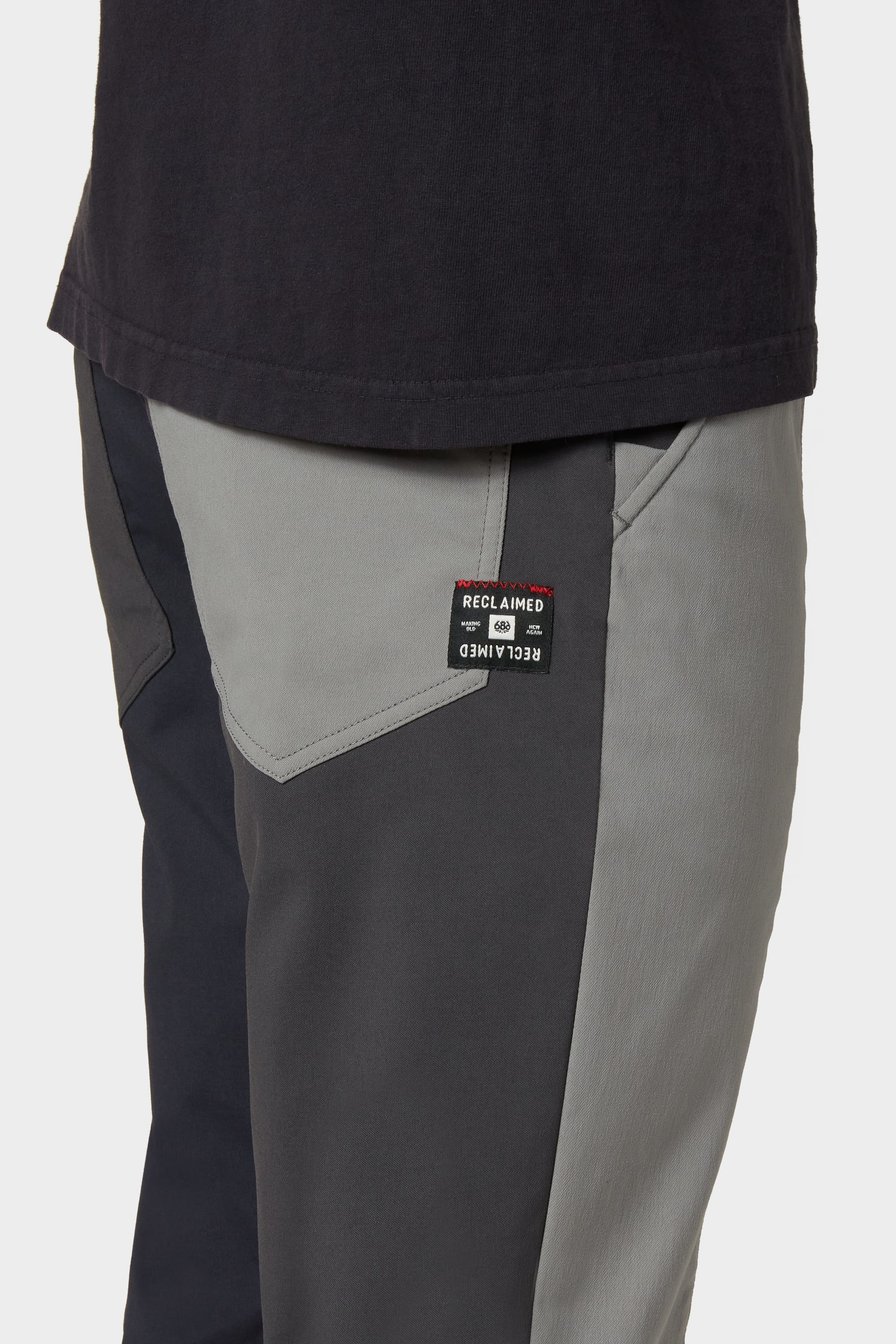 A close-up shows a person in the 686 Men's Everywhere® Reclaimed Pant - Slim Fit, color-blocked gray and black from deadstock fabrics. A black t-shirt peeks out at the waist, while a 686 Reclaimed label on the pocket highlights its eco-friendly design.