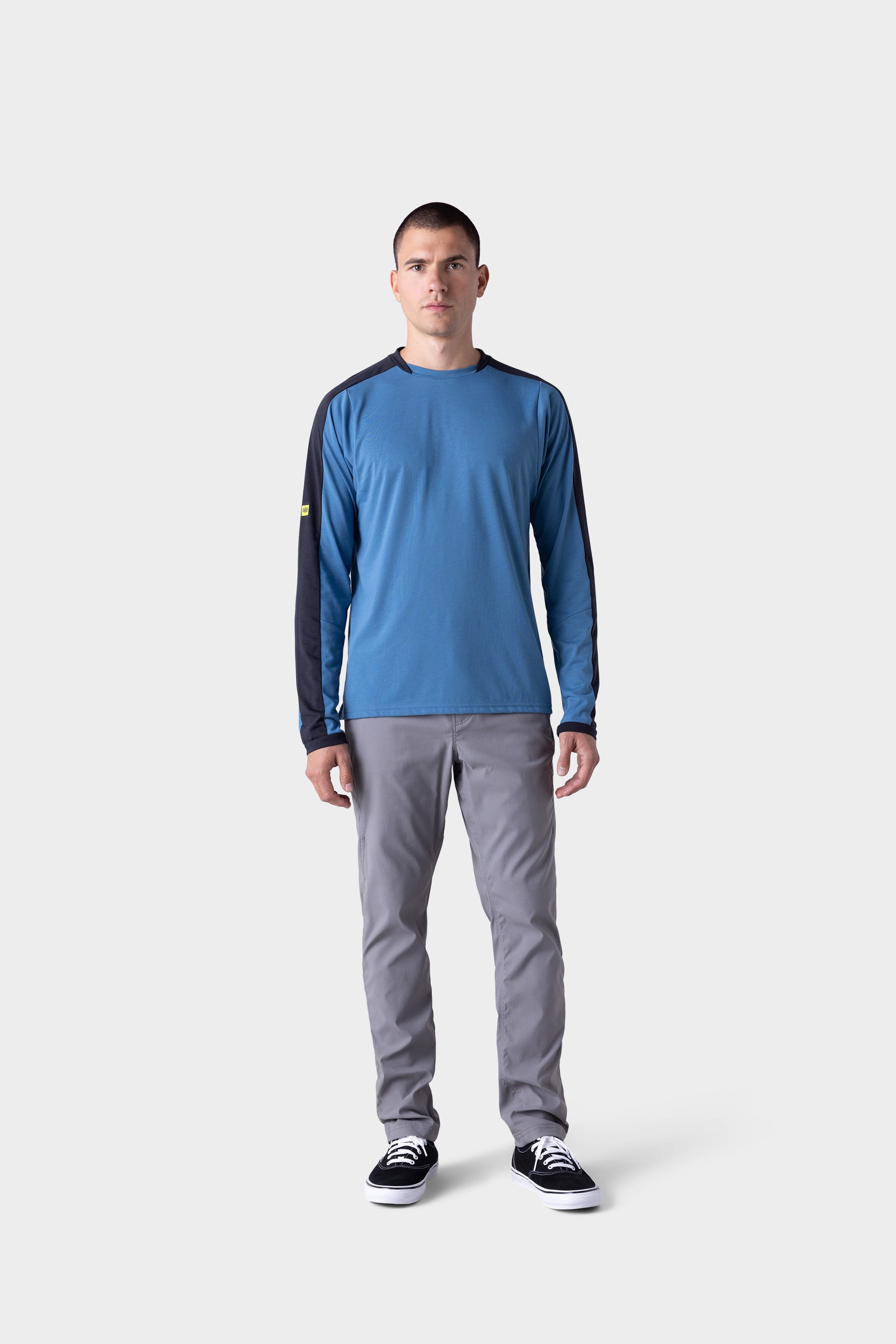 A man faces forward wearing the 686 Mens Rival Long Sleeve Bike Jersey by 686, paired with light gray pants and black-and-white sneakers, standing against a plain light gray background.