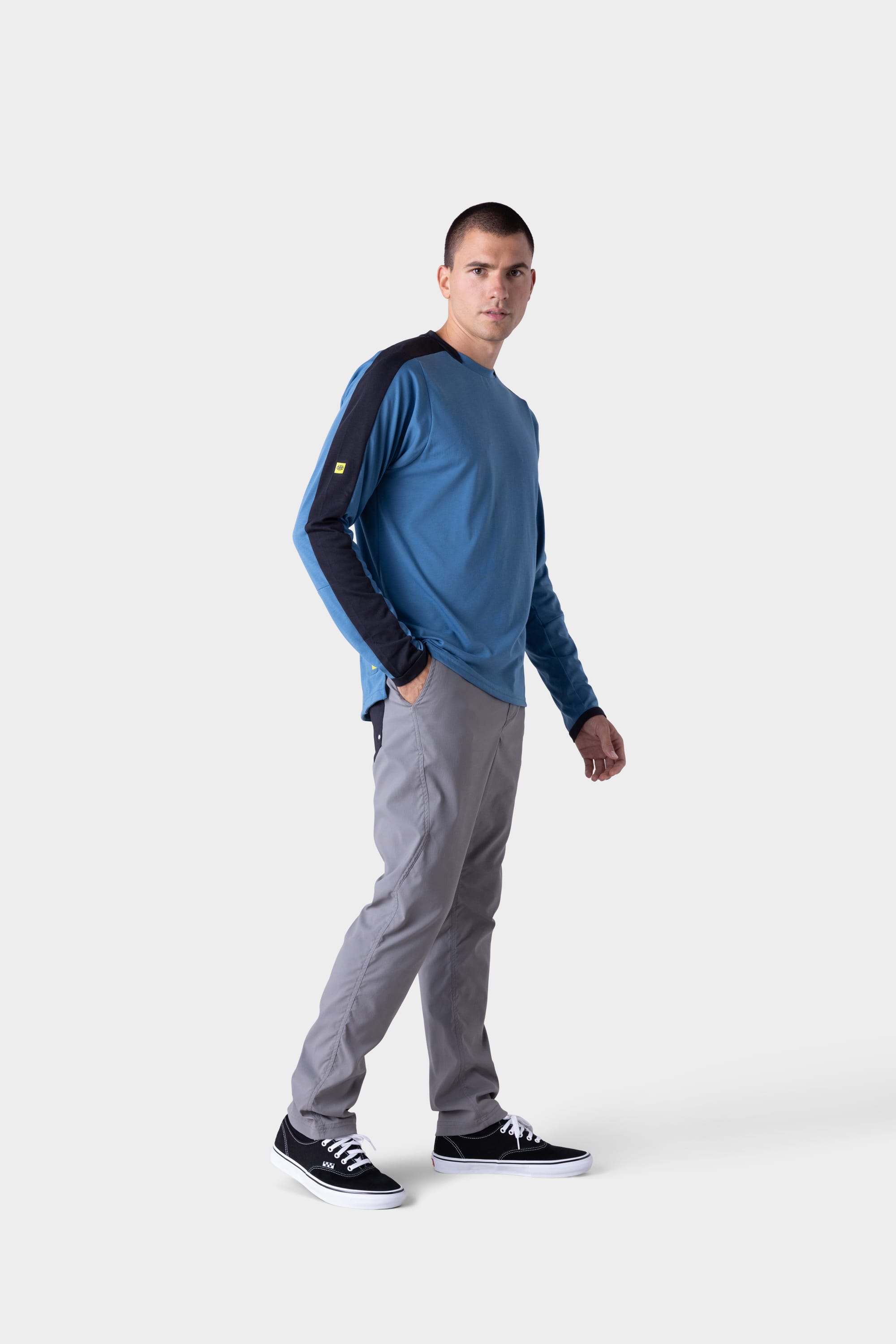 A man wears the 686 Mens Rival Long Sleeve Bike Jersey in blue with black accents, paired with gray pants and black sneakers, standing against a plain light gray background and looking slightly to his left.