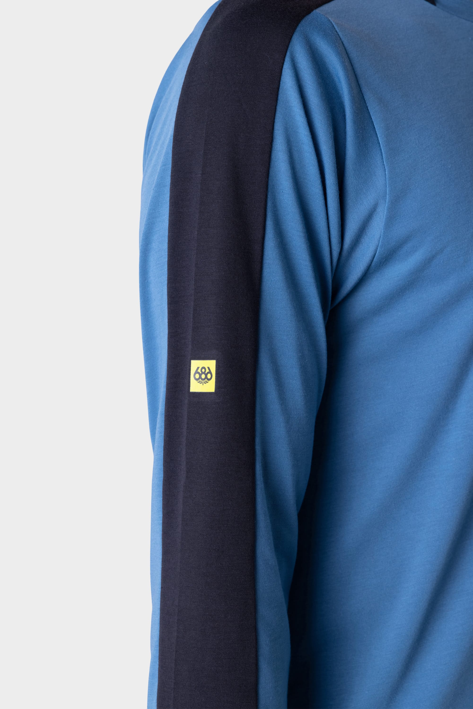 Close-up of a person wearing the 686 Mens Rival Long Sleeve Bike Jersey by 686, featuring a blue design with a thick black stripe along the shoulder and arm, plus a small yellow logo patch on the upper arm. The background is plain and light-colored.