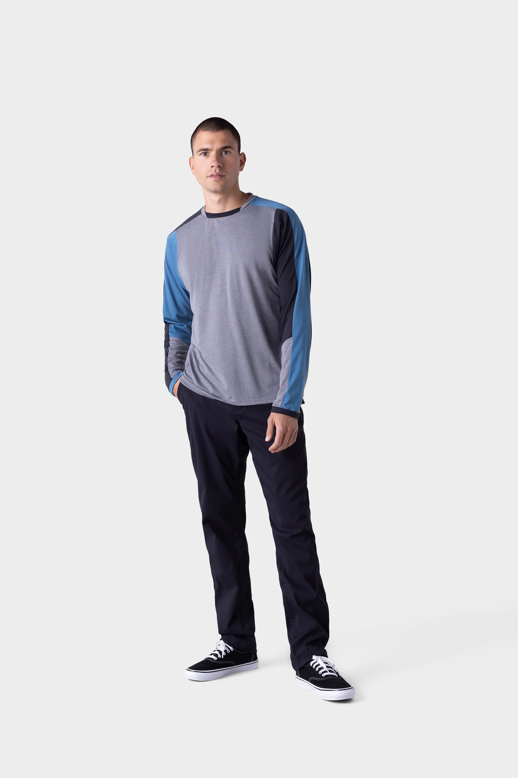 A man stands against a light gray background, wearing the 686 Mens Rival Long Sleeve Bike Jersey by 686. He pairs it with black pants and black sneakers with white soles, has his hands in his pockets, and looks straight ahead.