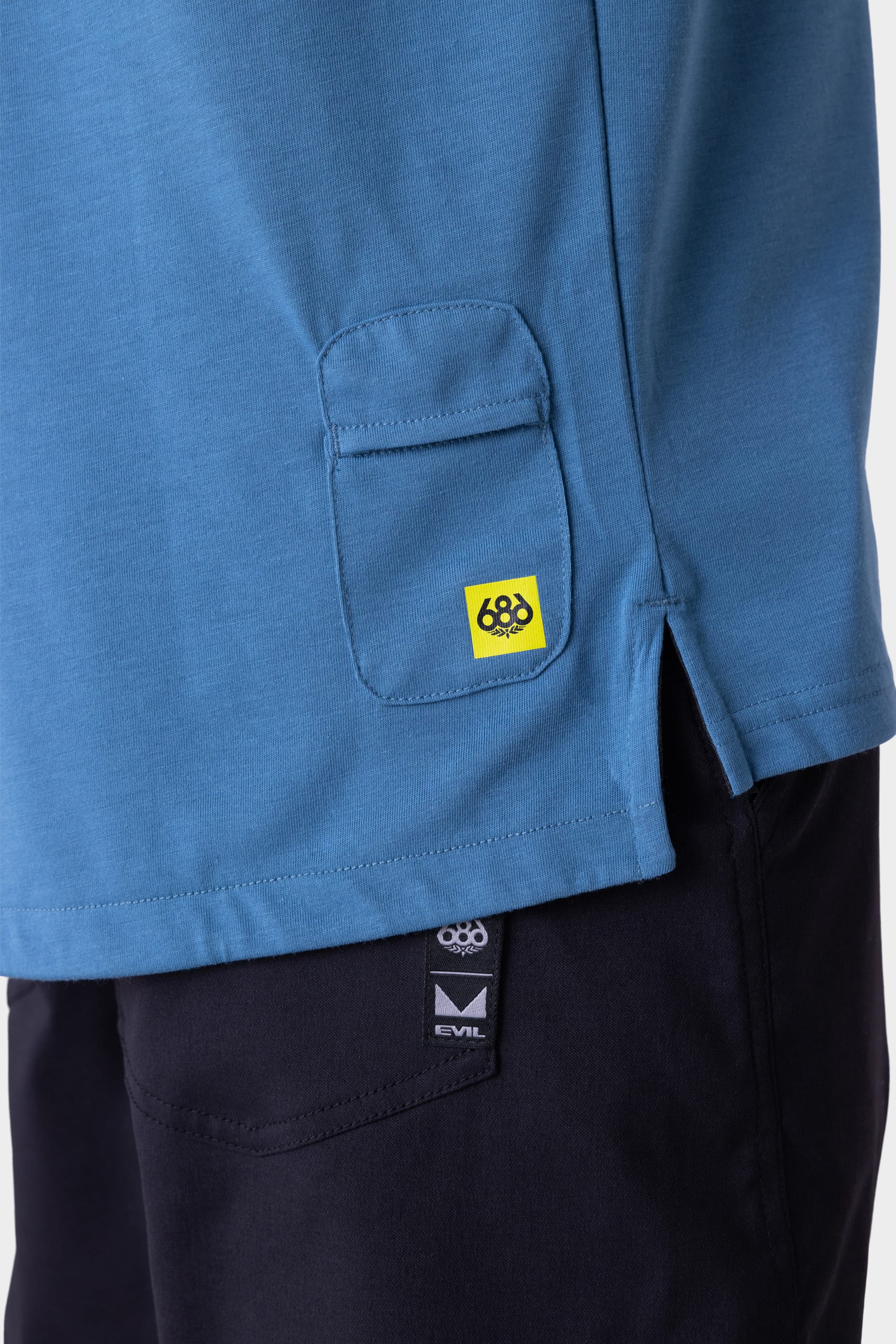 A close-up of a person wearing the 686 Mens Rival Short Sleeve Bike Jersey in blue, featuring a small flap pocket with a yellow 686 brand tag, paired with black pants showing visible 686 and EVIL brand tags near the waistband.