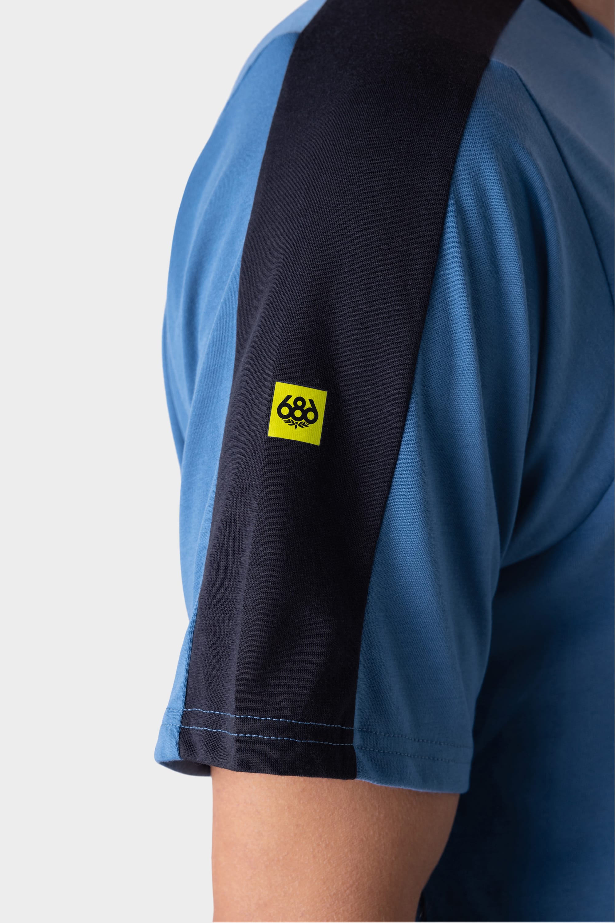 Close-up of a person wearing the 686 Mens Rival Short Sleeve Bike Jersey in blue, featuring a black shoulder stripe and a small yellow patch with the 686 logo. Their arm is visible, but their face is not shown.