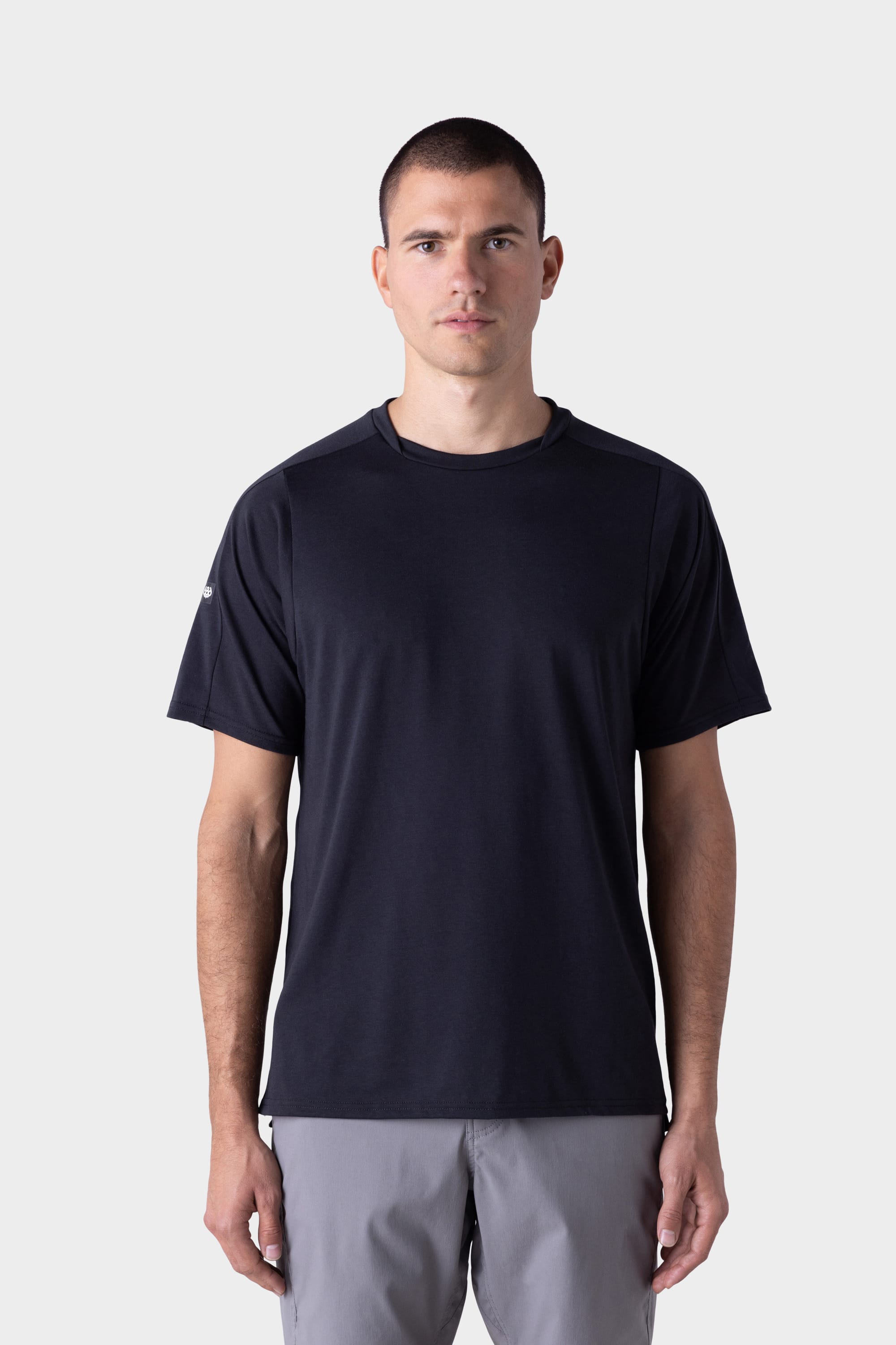 A man with short dark hair wears the 686 Mens Rival Short Sleeve Bike Jersey by 686, paired with light gray shorts, standing against a plain light gray background and facing forward with a neutral expression.