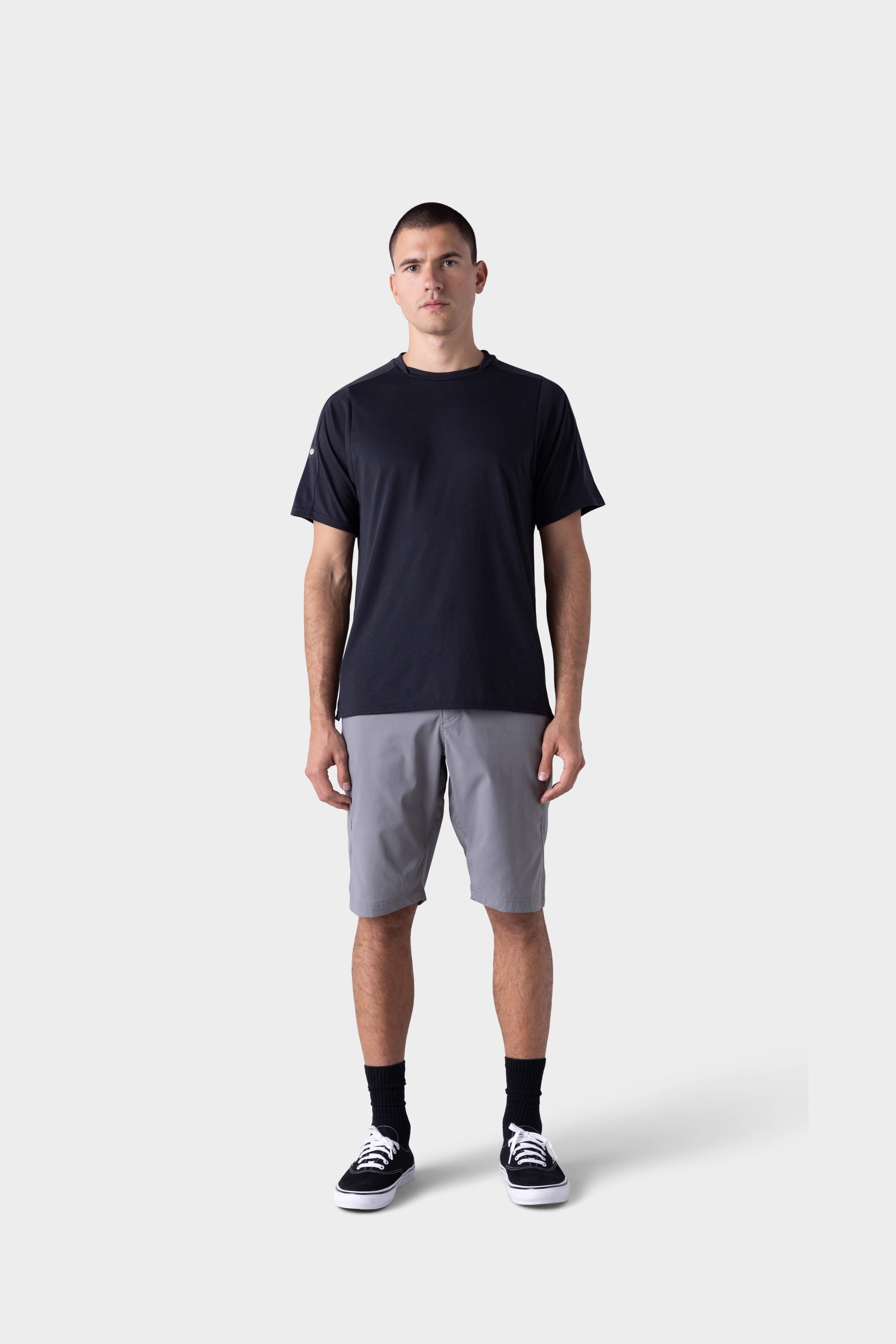 A man stands facing forward against a plain light gray background, wearing the 686 Mens Rival Short Sleeve Bike Jersey in black, paired with gray shorts, black socks, and black sneakers with white soles.