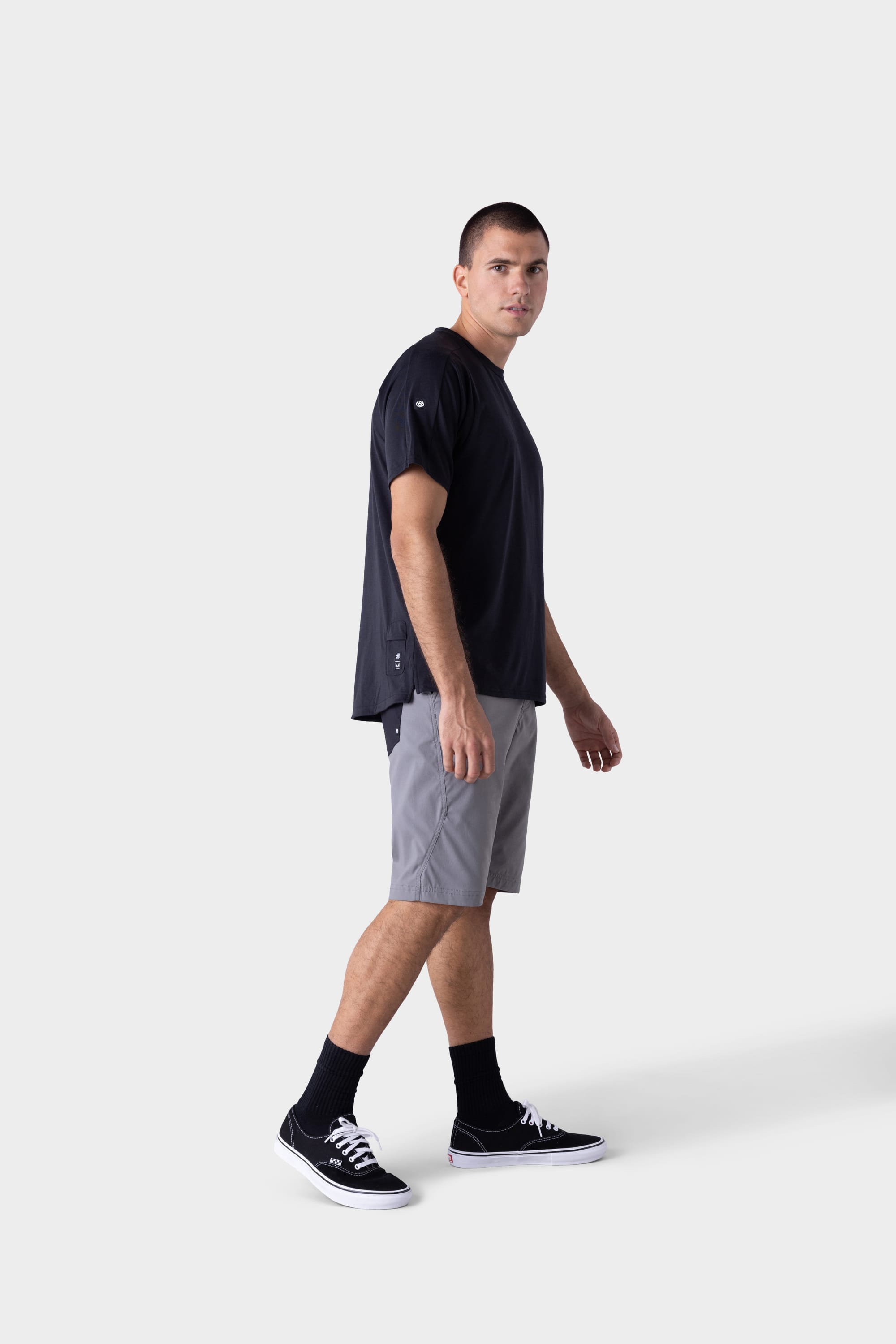 A man wearing the 686 Mens Rival Short Sleeve Bike Jersey stands against a plain light background, paired with light gray shorts, black socks, and black sneakers, looking slightly to the side.