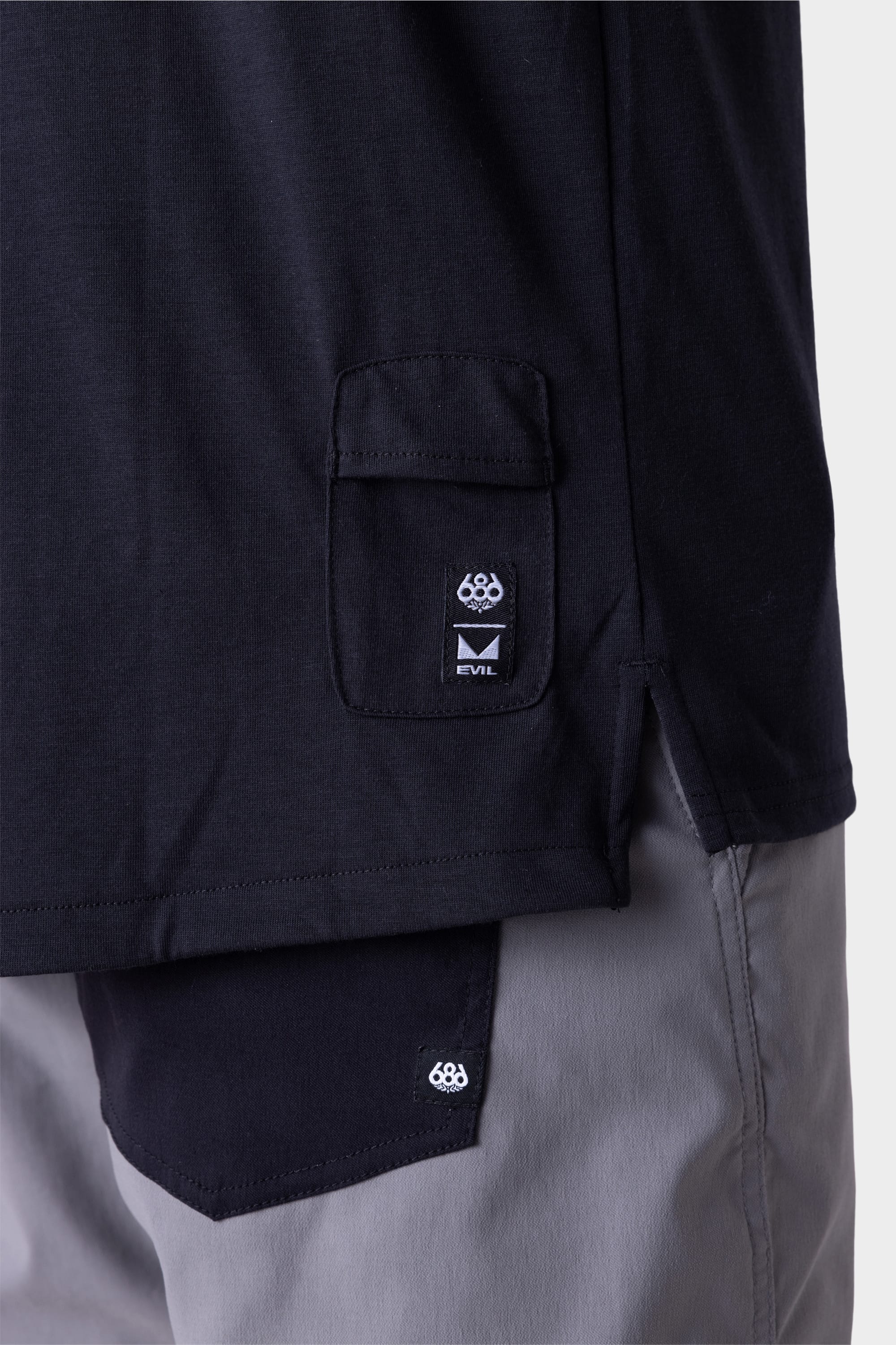 Close-up of a person wearing the 686 Mens Rival Short Sleeve Bike Jersey in black, featuring a flap pocket with the 686 logo label and EVIL. The light gray pants also display a matching 686 logo on the pocket.