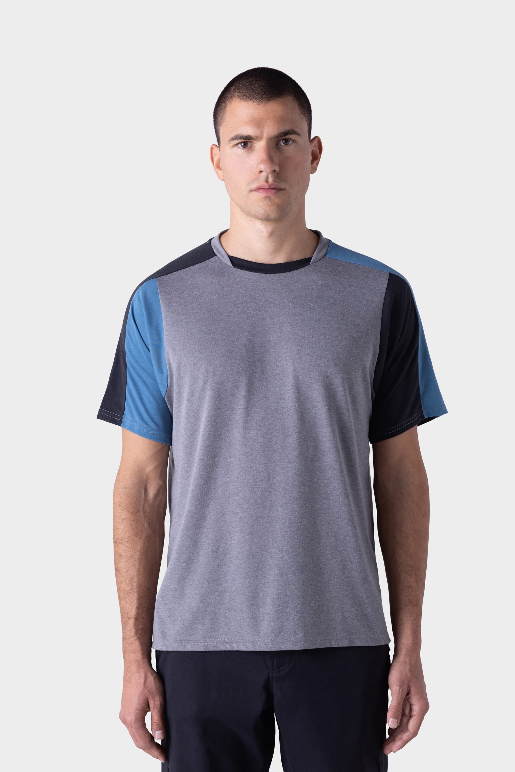 A man with short dark hair stands facing forward, wearing the 686 Mens Rival Short Sleeve Bike Jersey by 686, which features gray fabric with blue and black color-blocked sleeves, and dark pants, against a plain light background.