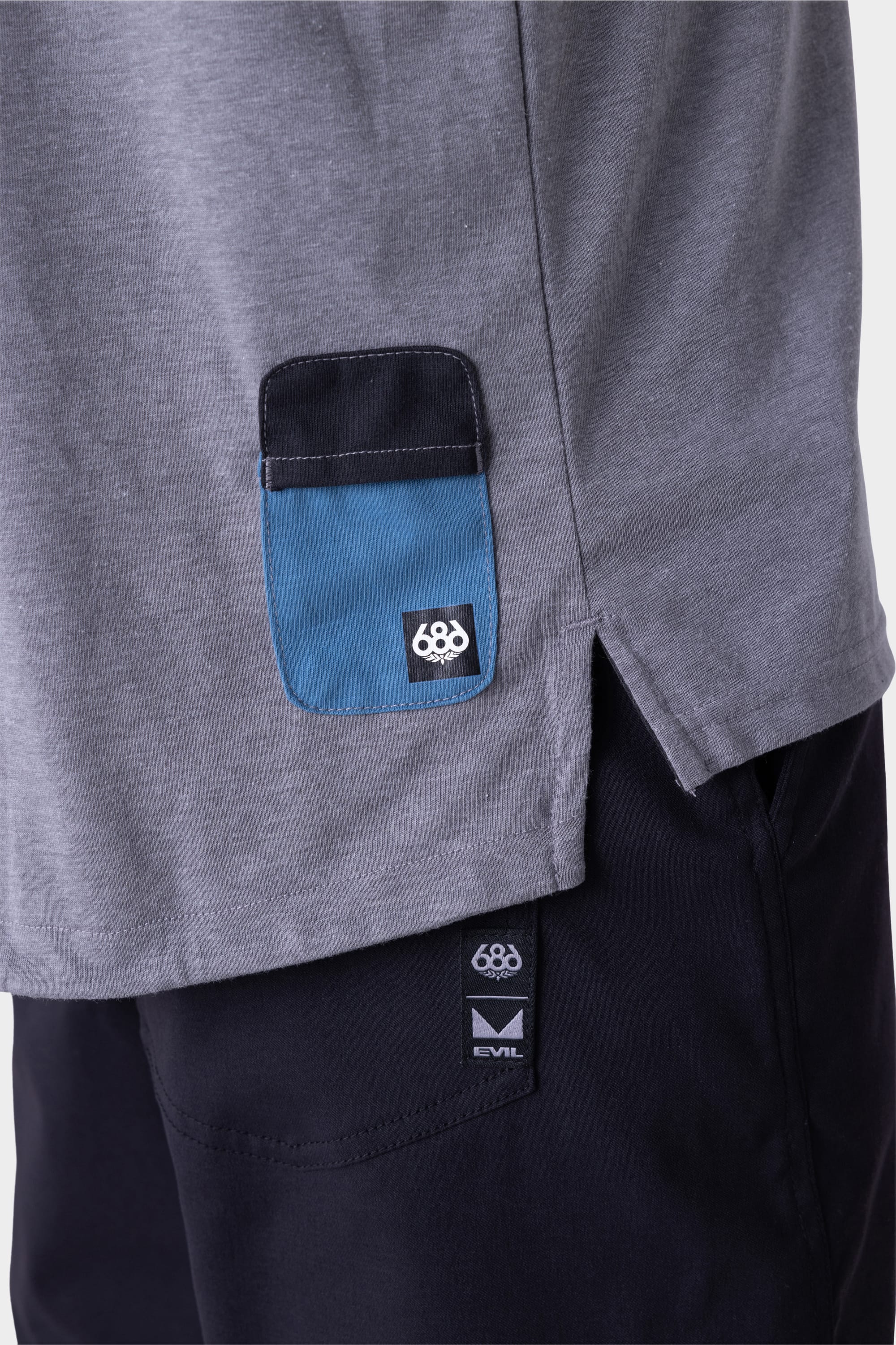Close-up of the 686 Mens Rival Short Sleeve Bike Jersey in gray with a small blue and black pocket featuring the 686 logo, paired with black pants displaying a visible 686 EVIL tag near the waistband.