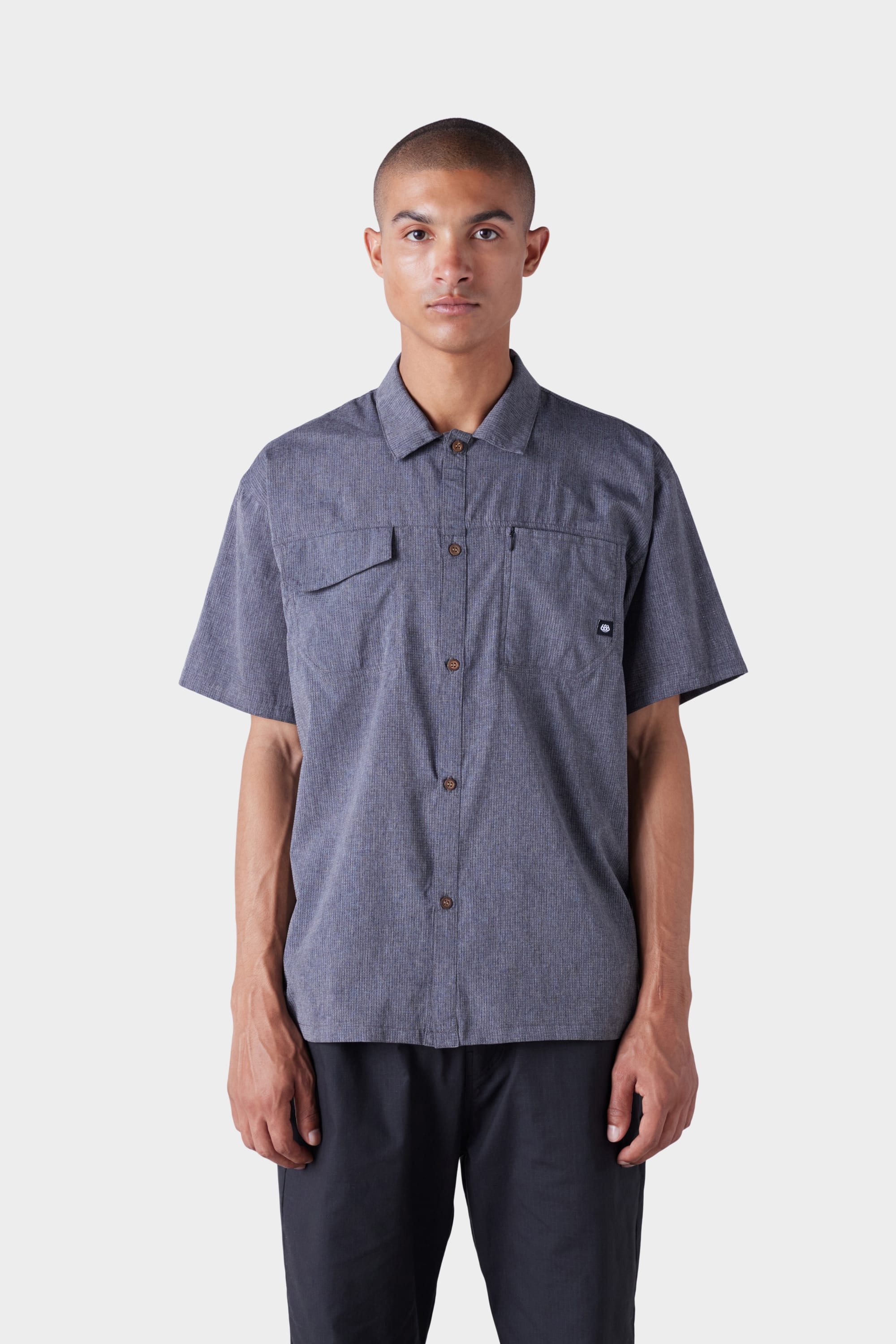 A person faces forward against a white background, wearing the 686 Mens Seeker Perforated Button Up by 686, a short-sleeve gray shirt with two chest pockets, paired with black pants.