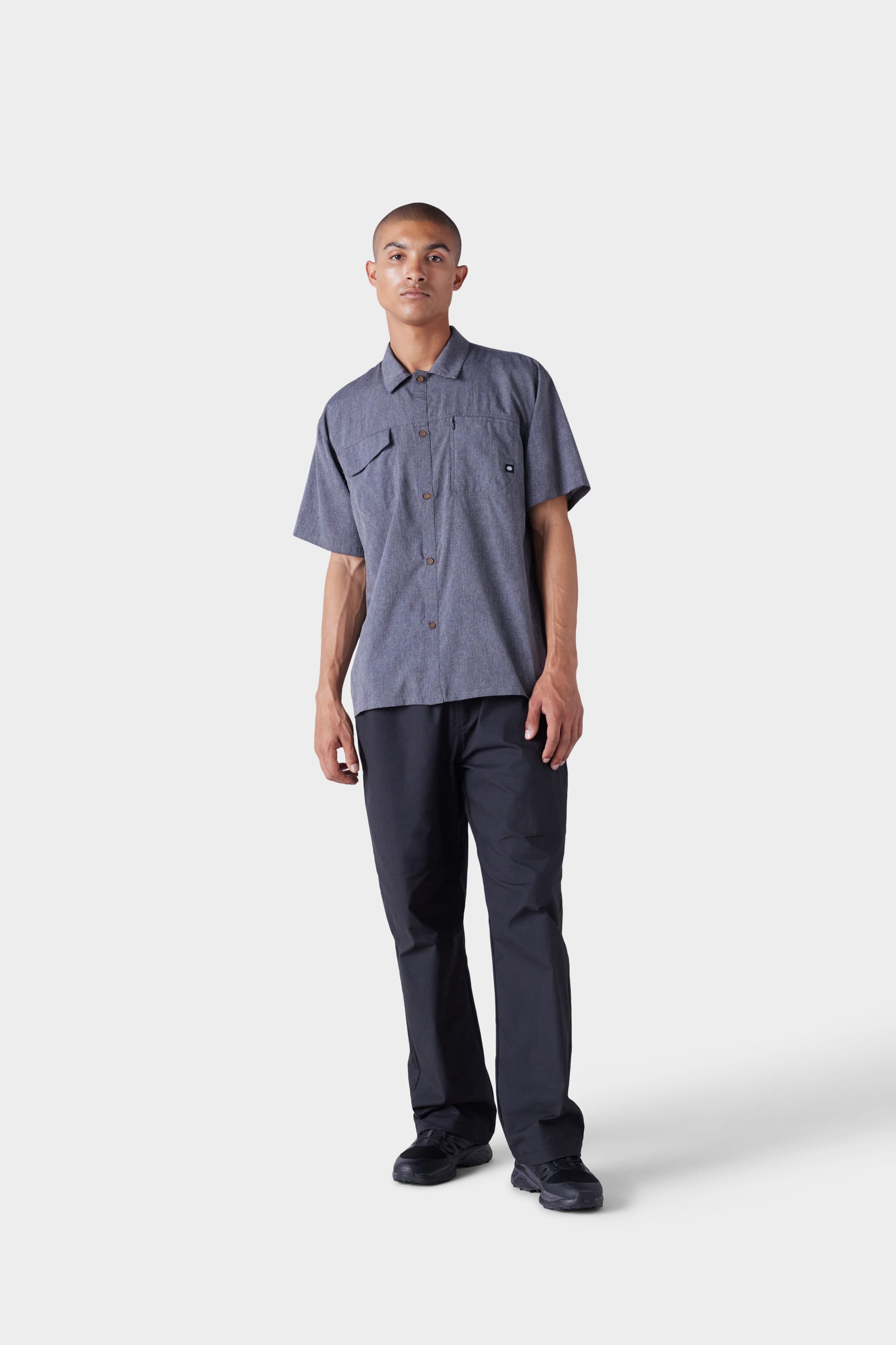 A person with short hair and a neutral expression wears the 686 Mens Seeker Perforated Button Up gray shirt by 686, featuring two chest pockets, paired with black pants and black shoes against a plain white background.