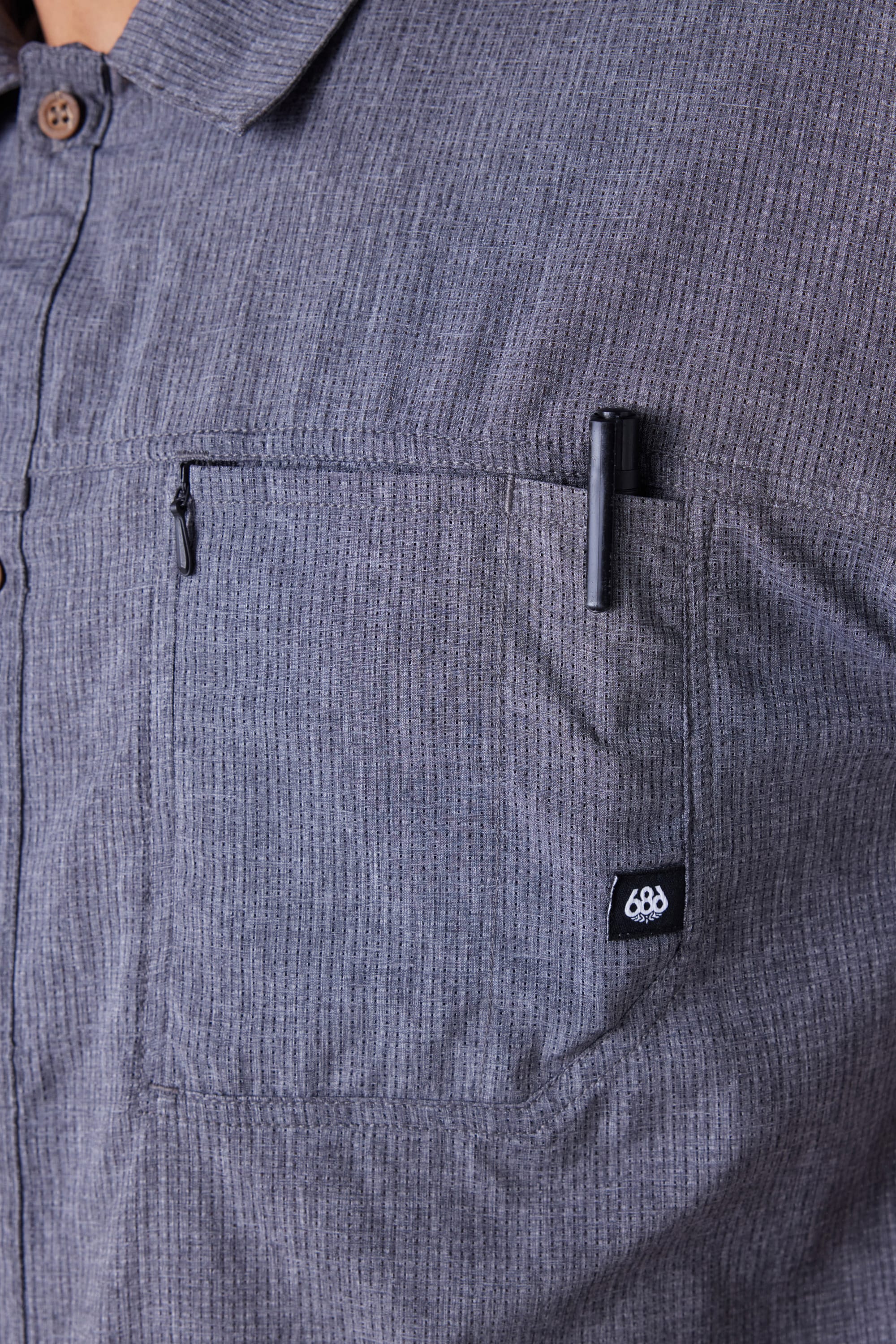 A close-up of the 686 Mens Seeker Perforated Button Up shows its grey vertical stripes, front pocket with a black pen tucked inside, and a small black tag featuring the white 686 logo on the pocket.