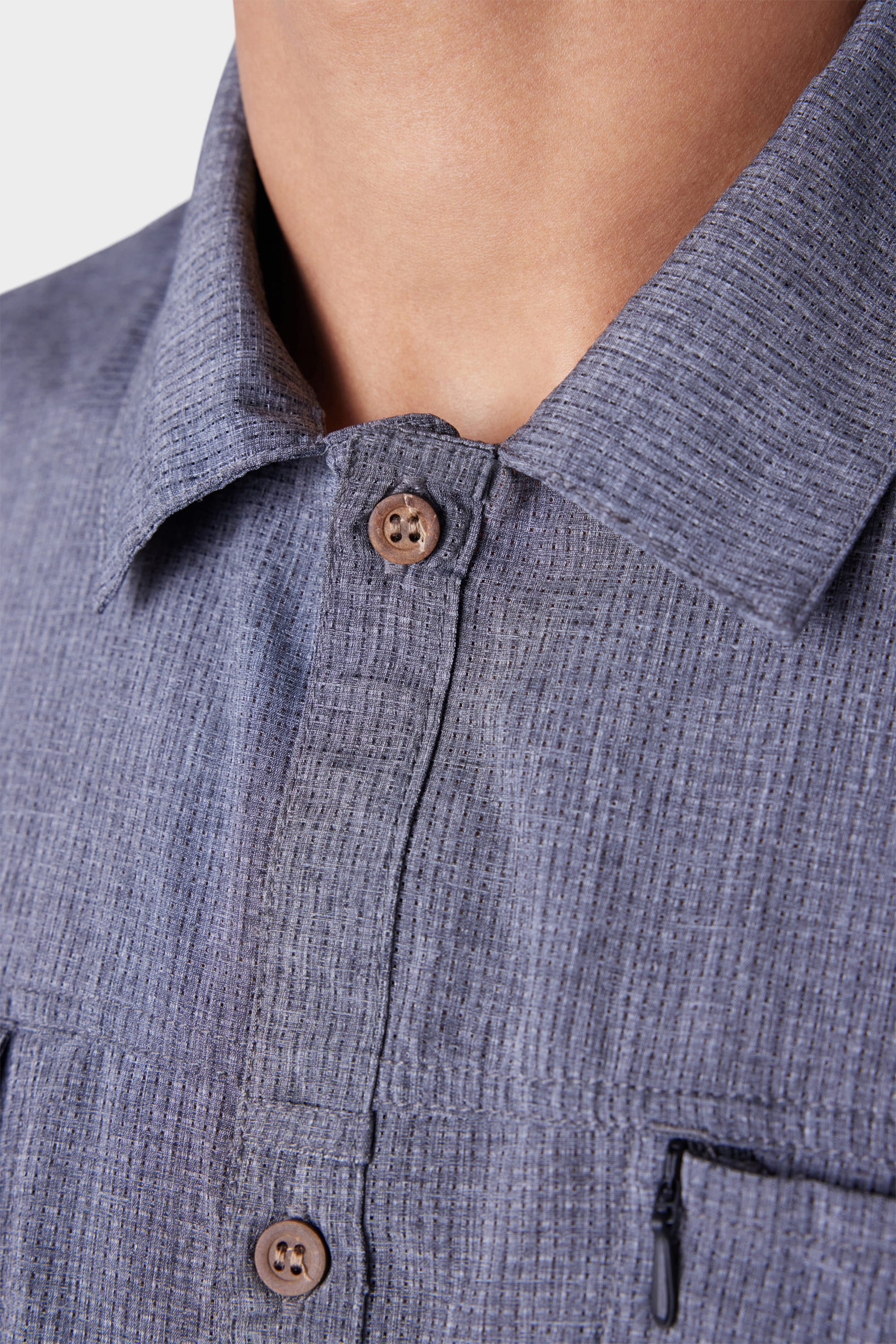 Close-up of a person wearing the 686 Mens Seeker Perforated Button Up, a textured blue-gray collared shirt by 686 with brown buttons and a visible vertical zipper pocket. The background is plain white.