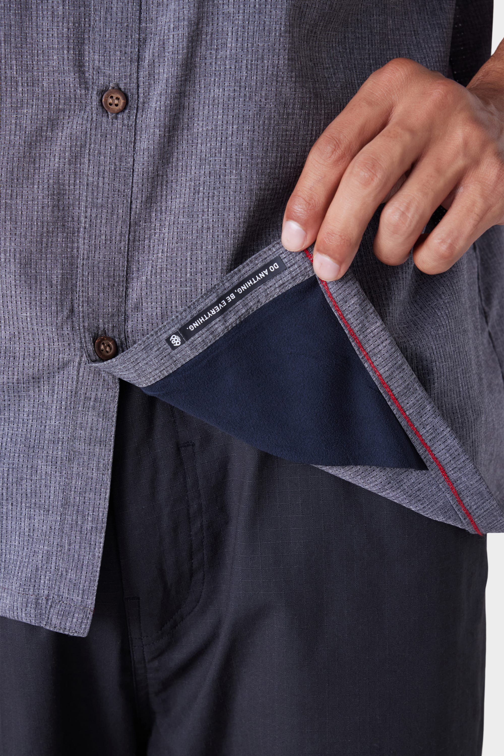 Someone lifts the bottom edge of the 686 Mens Seeker Perforated Button Up by 686, showing a hidden interior pocket lined with dark fabric and a red seam. They are wearing black pants.