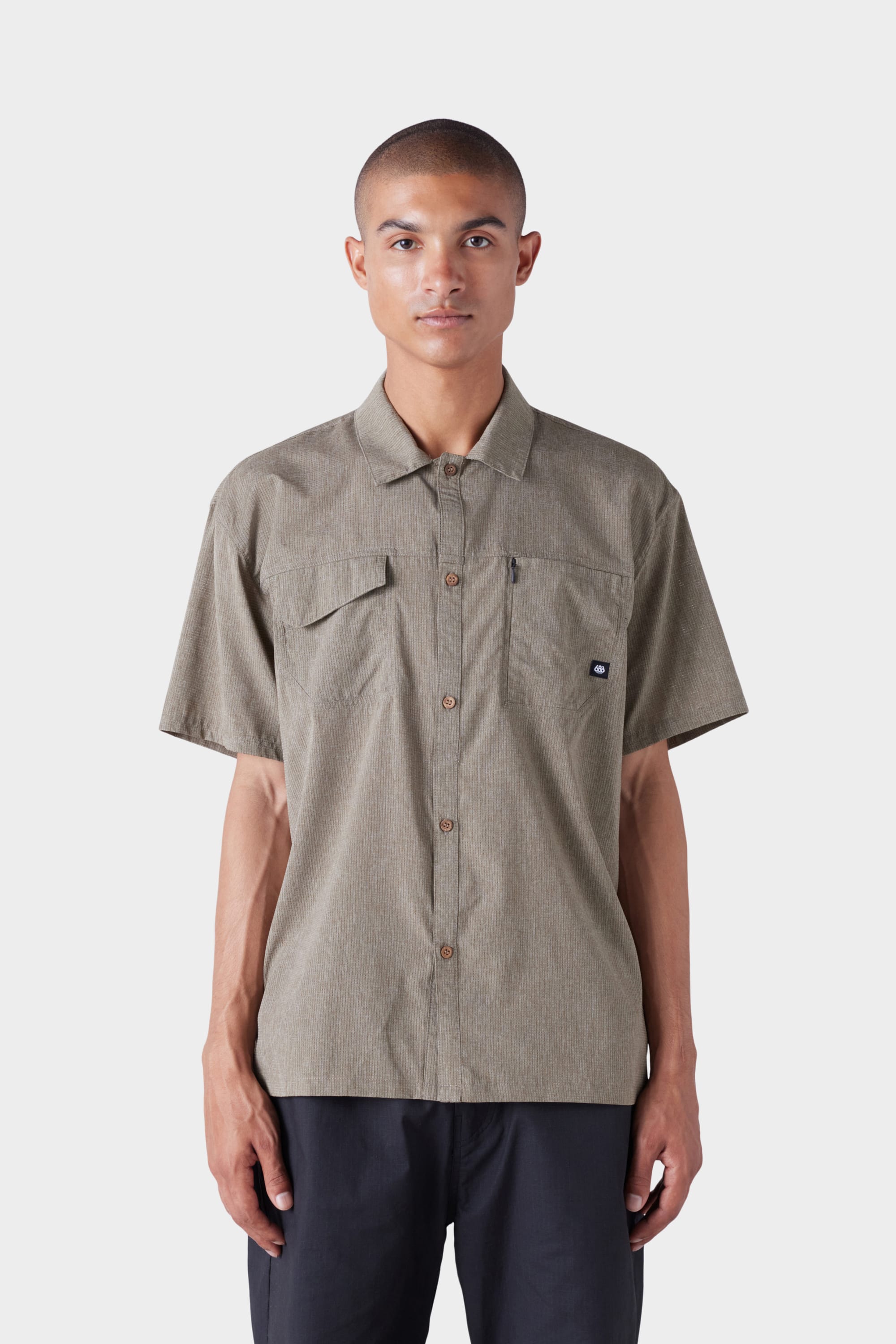 A man with short hair wears the 686 Mens Seeker Perforated Button Up, a short-sleeved olive-green shirt with a chest pocket, paired with black pants, standing against a plain white background.