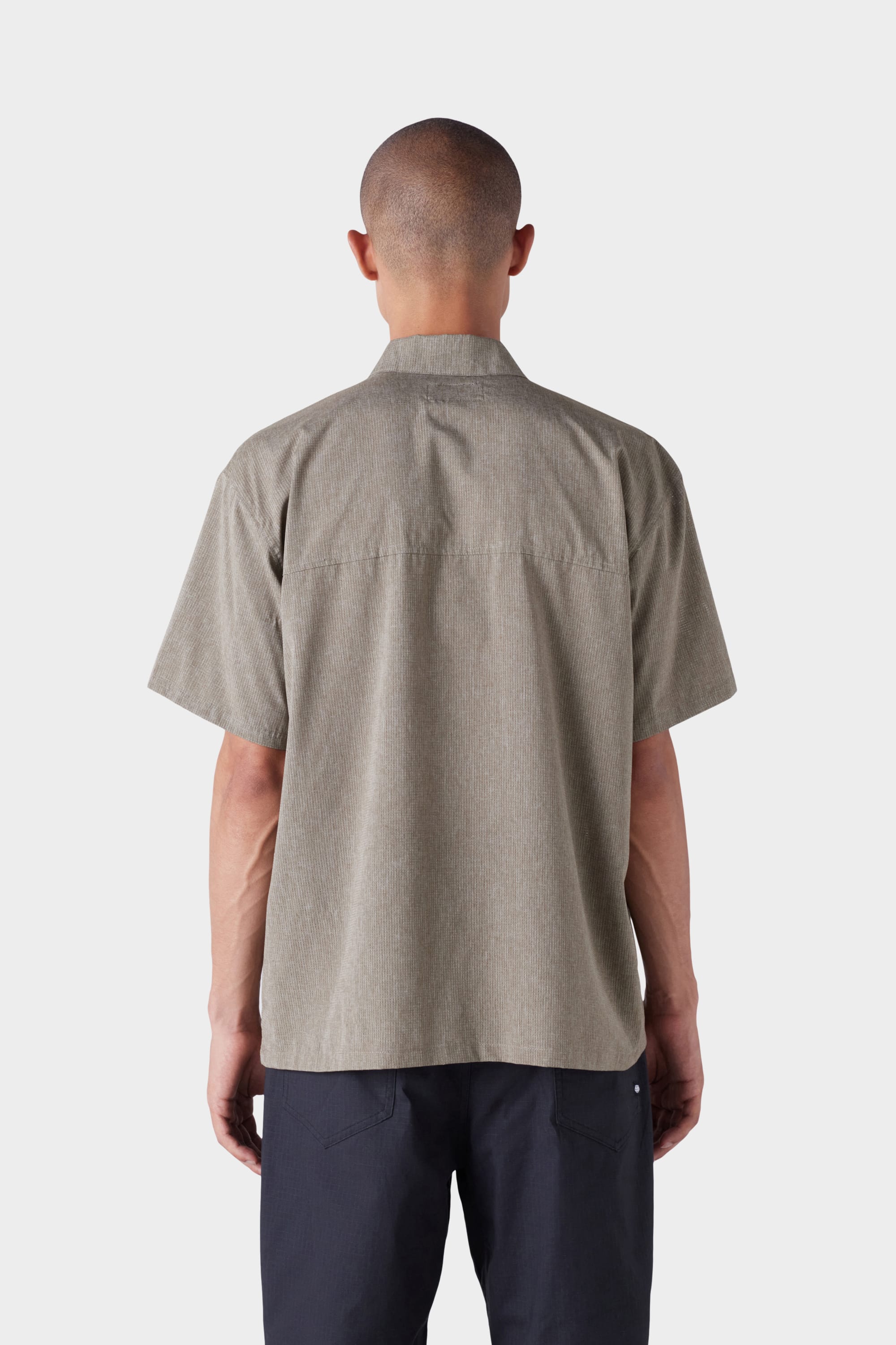 A person with closely cropped hair is shown from behind, wearing the 686 Mens Seeker Perforated Button Up in light brown and dark pants against a plain white background.