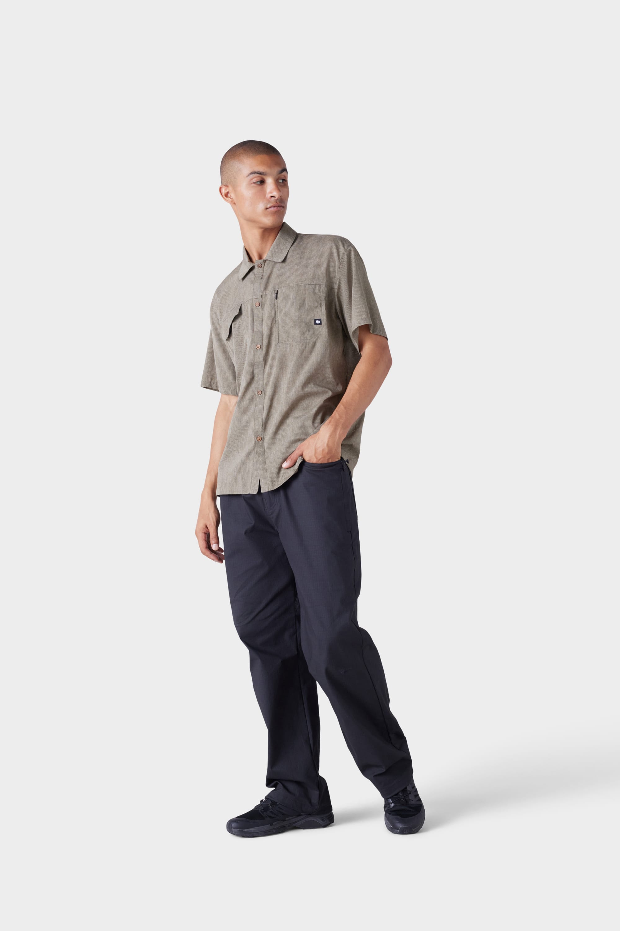 A person with short hair wears the 686 Mens Seeker Perforated Button Up shirt and dark pants with black shoes, standing and looking to the side against a plain light background.