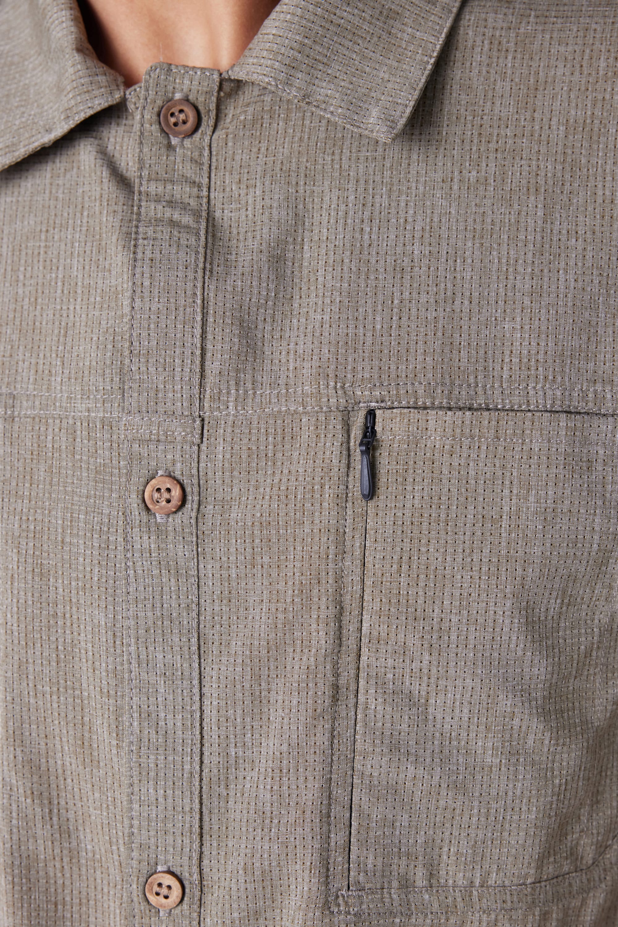 Close-up of the 686 Mens Seeker Perforated Button Up, a light brown collared shirt with vertical pinstripes, chest zipper pocket, and button front. Only the lower face and upper chest of the model are visible. Brand: 686.
