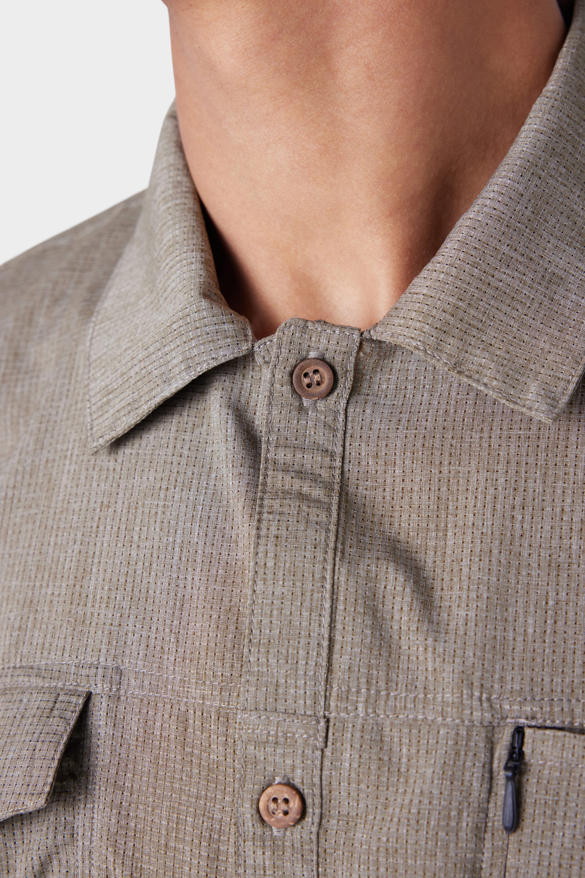 Close-up of a person wearing the 686 Mens Seeker Perforated Button Up, showing the light brown textured fabric, collared neckline, two visible brown buttons, and part of a zippered pocket. Only the neck and upper chest are shown.