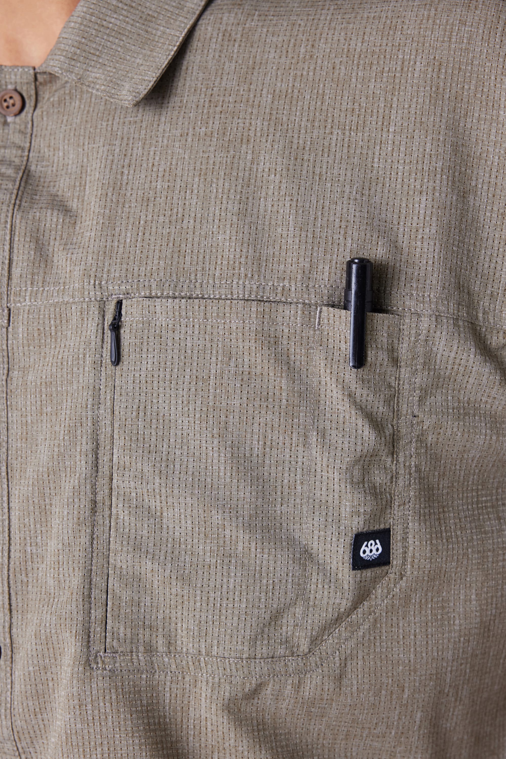 Close-up of the 686 Mens Seeker Perforated Button Up shirt in beige, featuring a chest pocket with a vertical zipper, a black pen clipped to the pocket, and a small black 686 tag sewn on the pockets edge.